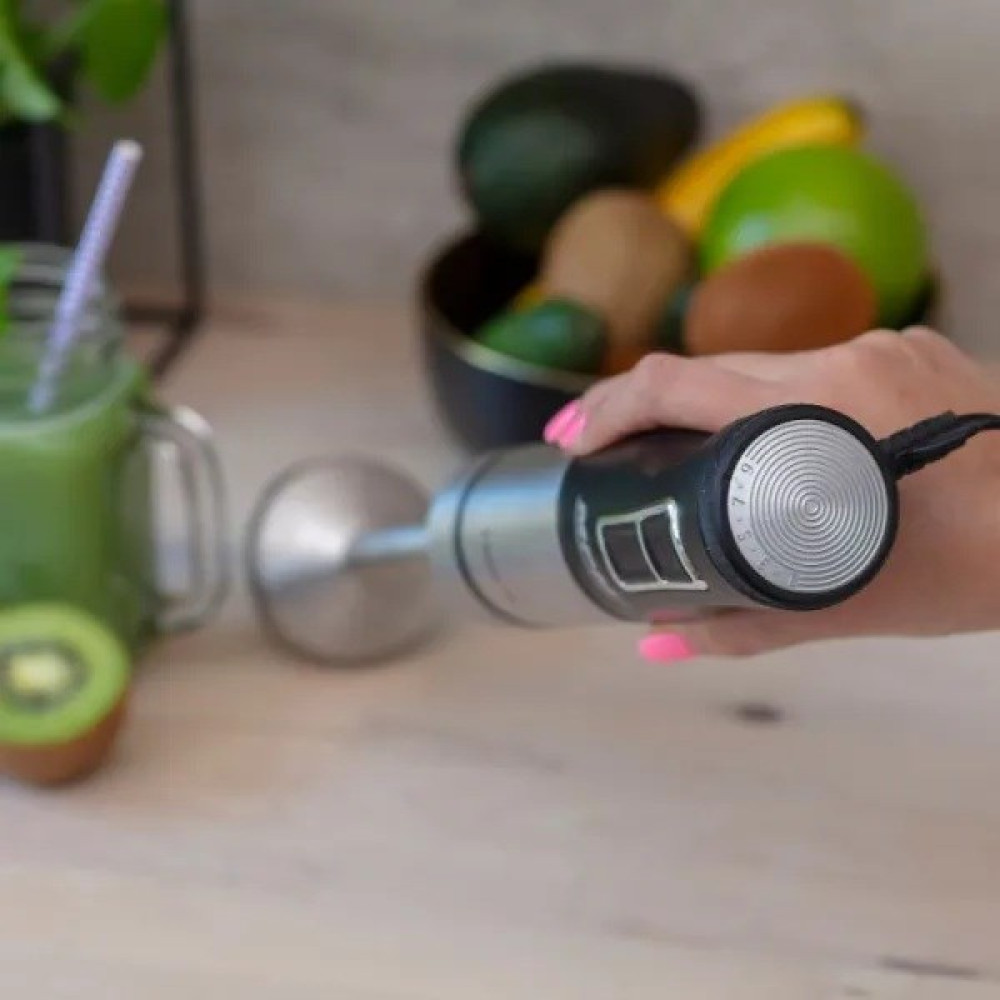 Una mano tiene un frullatore a immersione vicino a un bicchiere di frullato e ciotola di frutta.