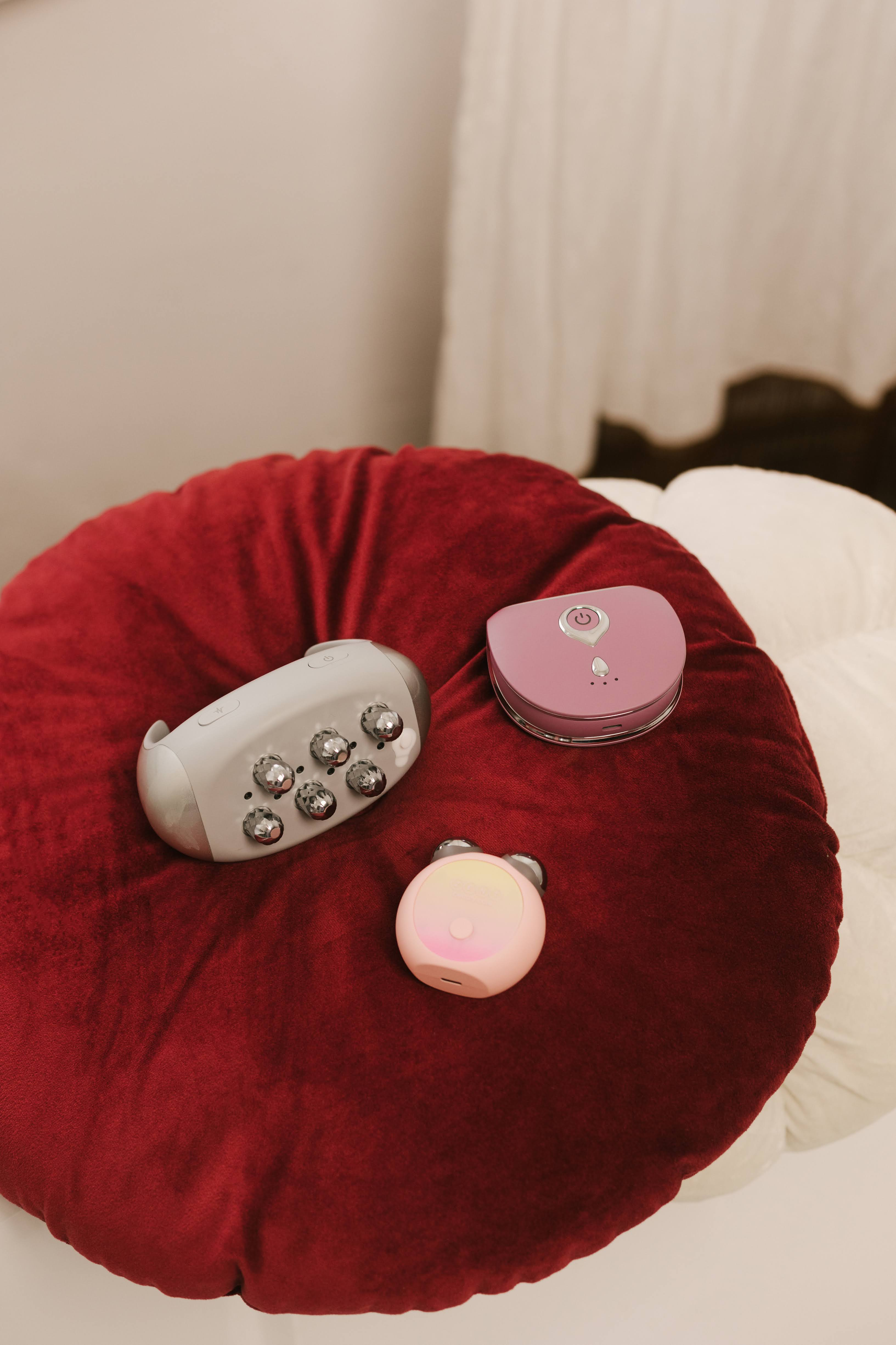 Trzy urządzenia kosmetyczne na czerwonej poduszce: jeden szary, dwa różowe.