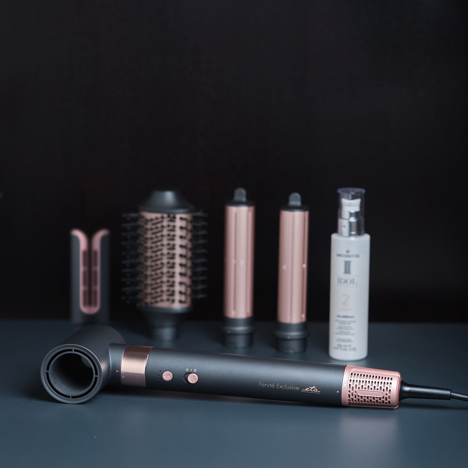 A dark hair dryer with rose gold attachments and a bottle on a table.