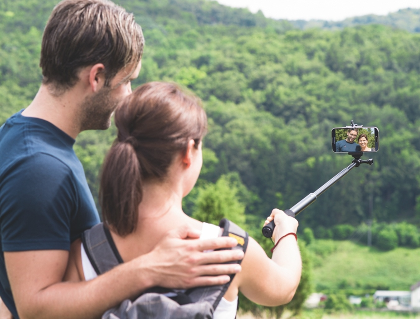 Una coppia scatta un selfie con un selfie stick davanti a un paesaggio collinare verde.