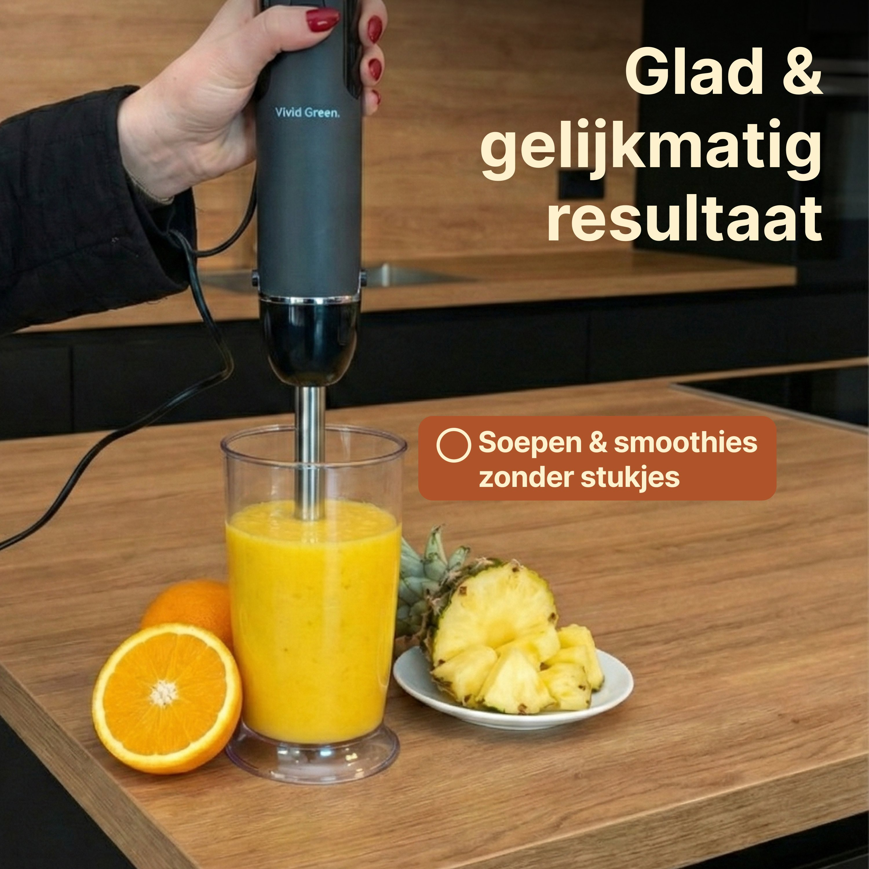 A hand blender mixes a yellow smoothie in a glass with oranges and pineapple beside it.
