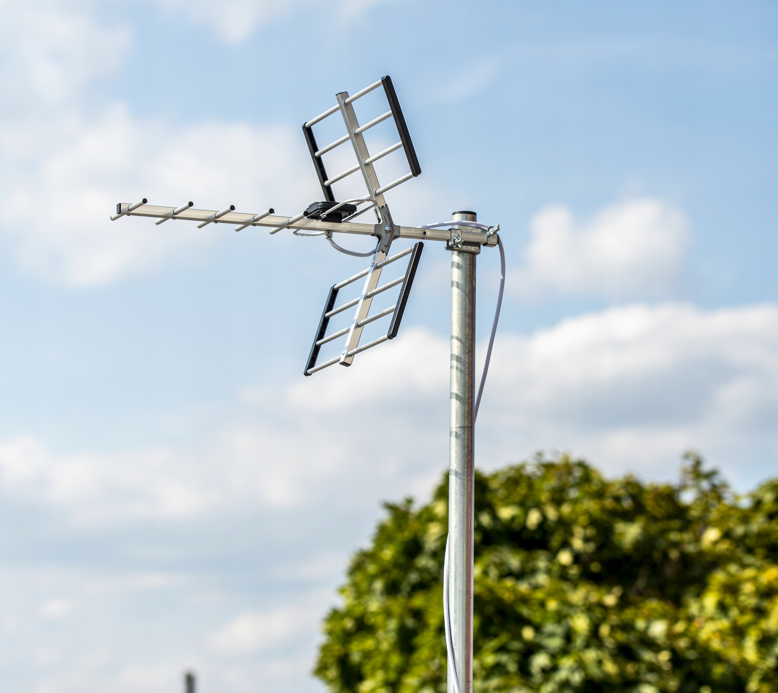 Eine silberfarbene Antenne steht vor einem hellblauen Himmel mit weißen Wolken, mit grünem Laub im Hintergrund.