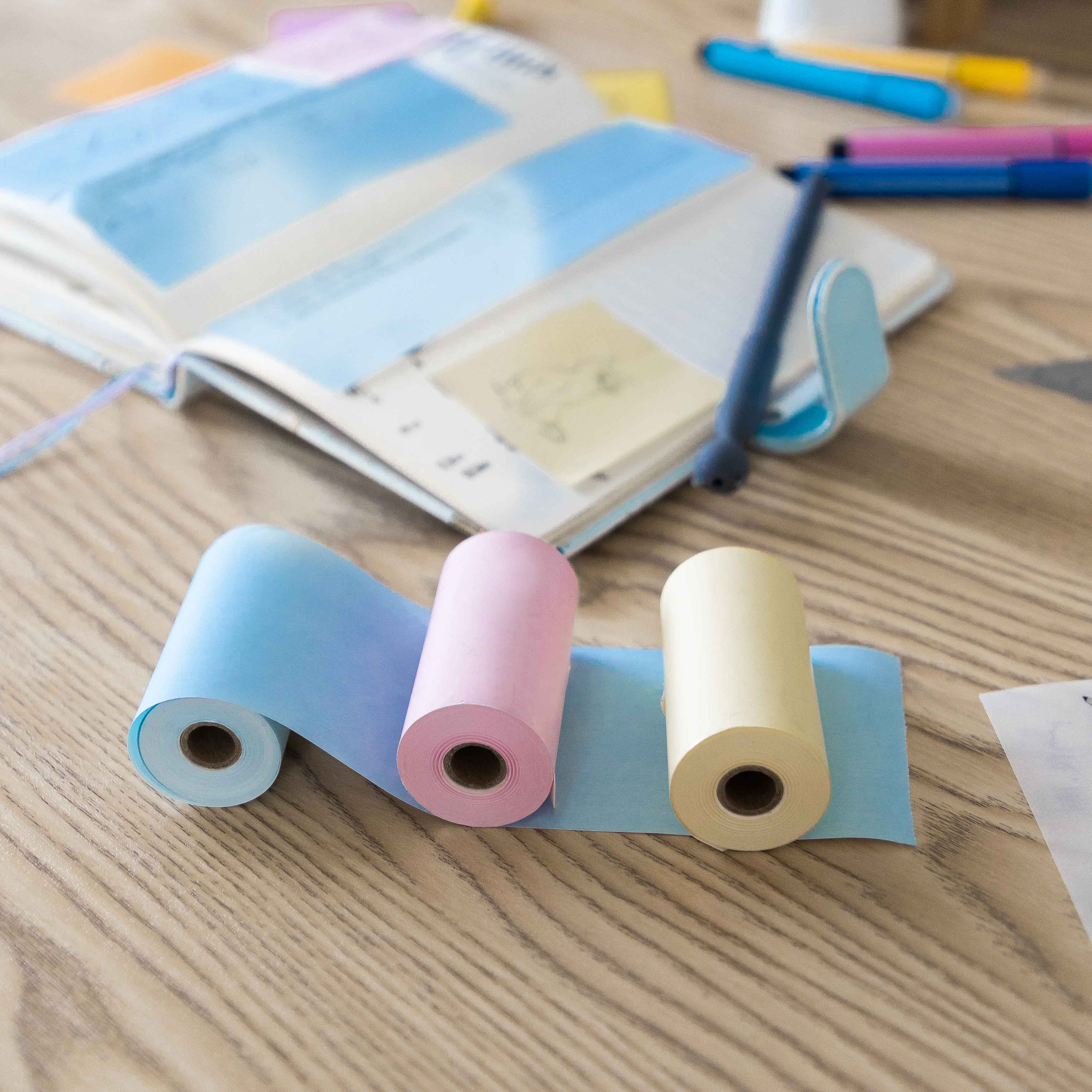 Drei Rollen farbiges Thermopapier (blau, rosa, gelb) auf einem Holzschreibtisch mit offenem Planer und Stiften.