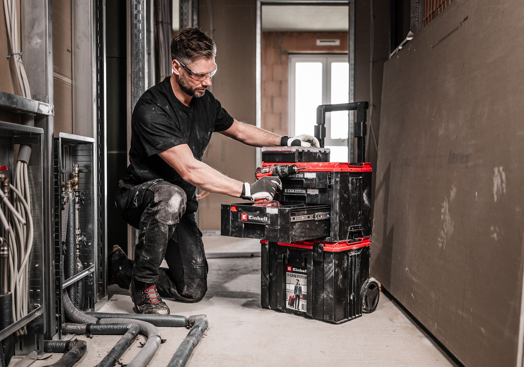 Ein Mann mit Schutzbrille kniet und sortiert Werkzeuge in einem schwarz-roten Werkzeugkastensystem auf einer Baustelle.