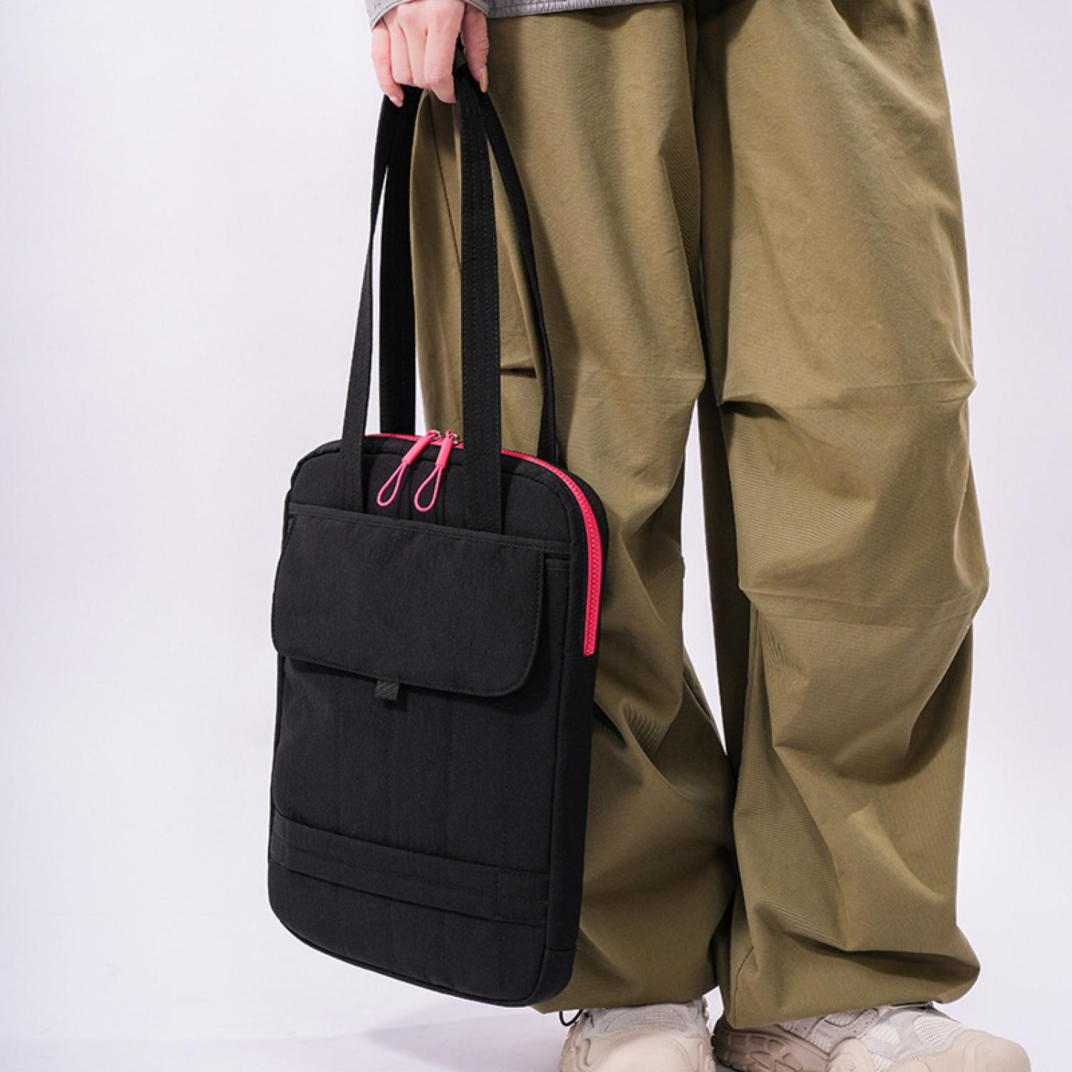 Person hält schwarze Tasche mit pinkem Reißverschluss. Grüne Hose, weiße Schuhe.