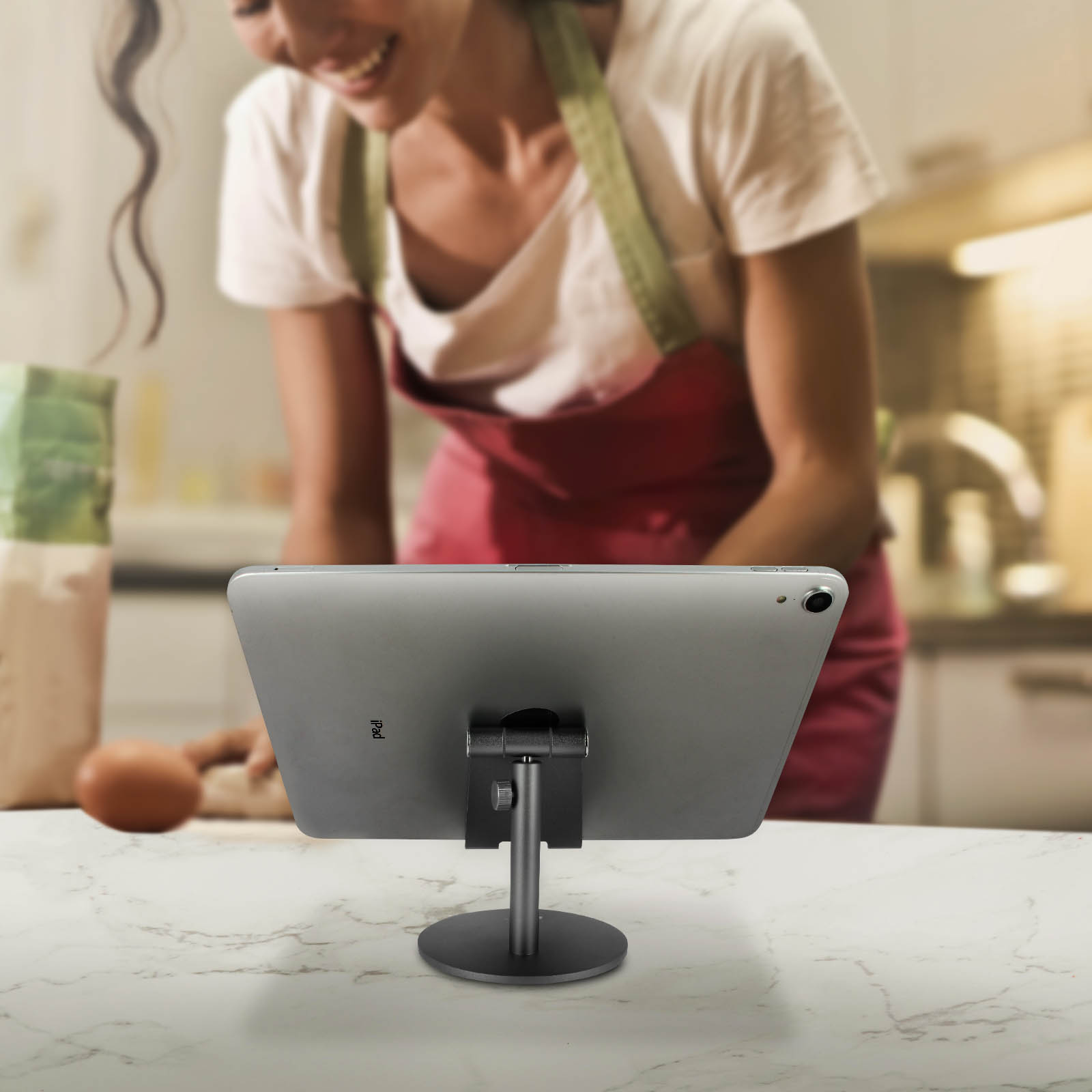 Un tablet su un supporto con una donna che cucina sullo sfondo.