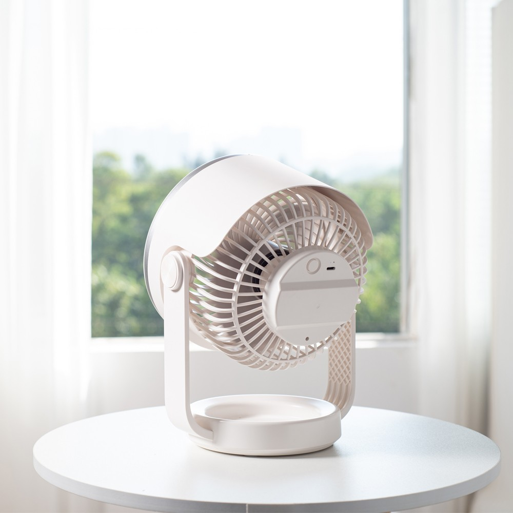 Un ventilador blanco está sobre una mesa redonda frente a una ventana.