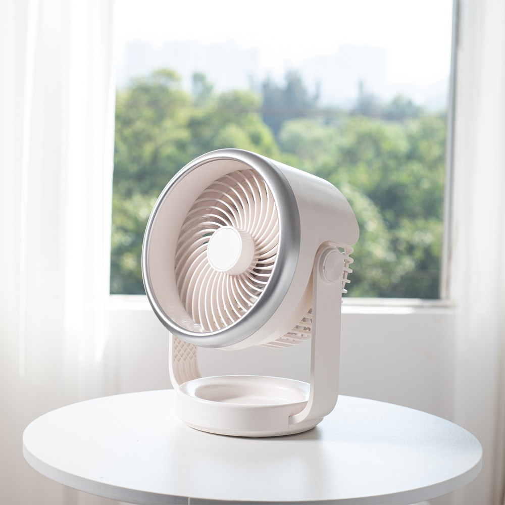 Un ventilador de mesa blanco está sobre una mesa redonda frente a una ventana con árboles.