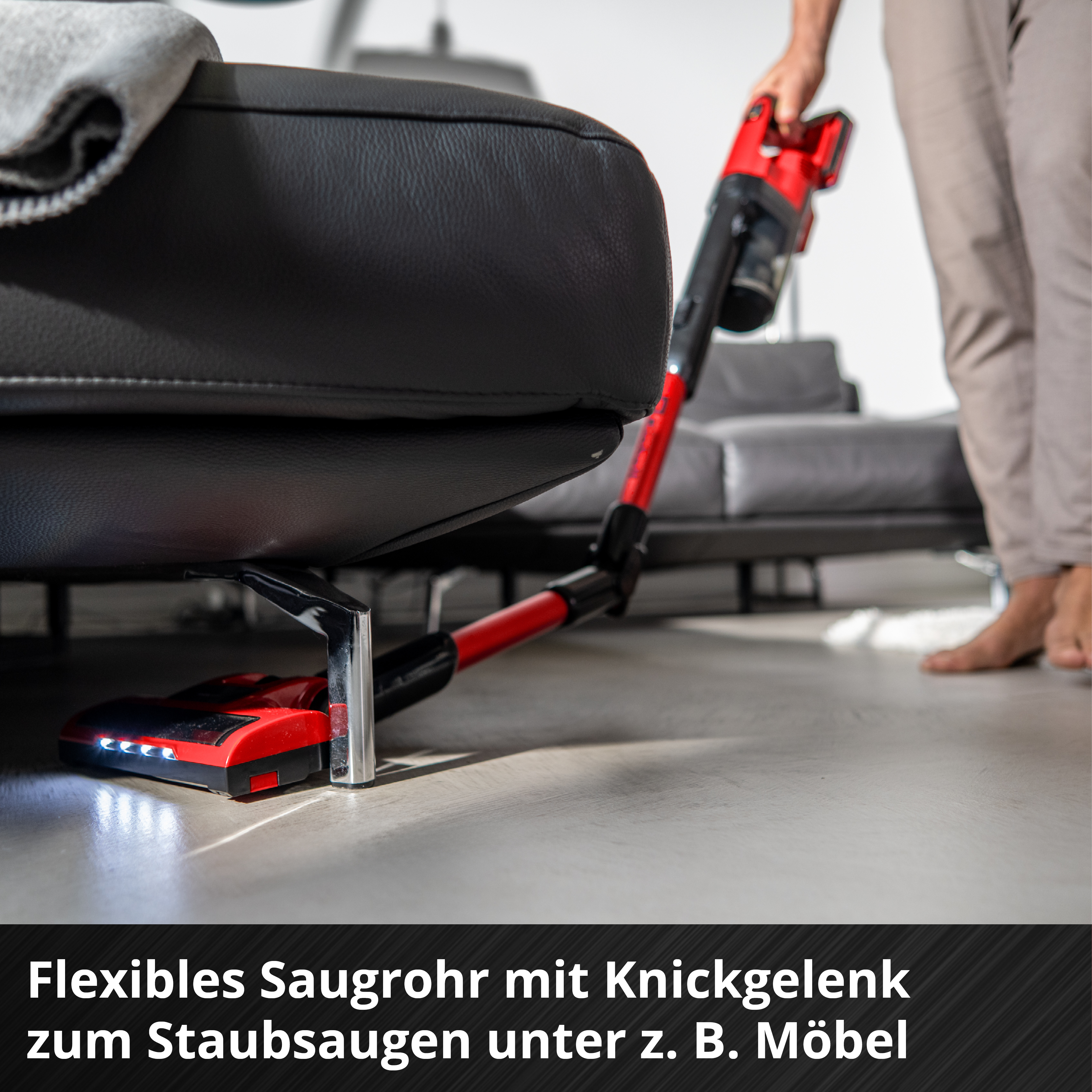 Ein roter Handstaubsauger mit LED-Lichtern reinigt unter einem schwarzen Ledersofa.