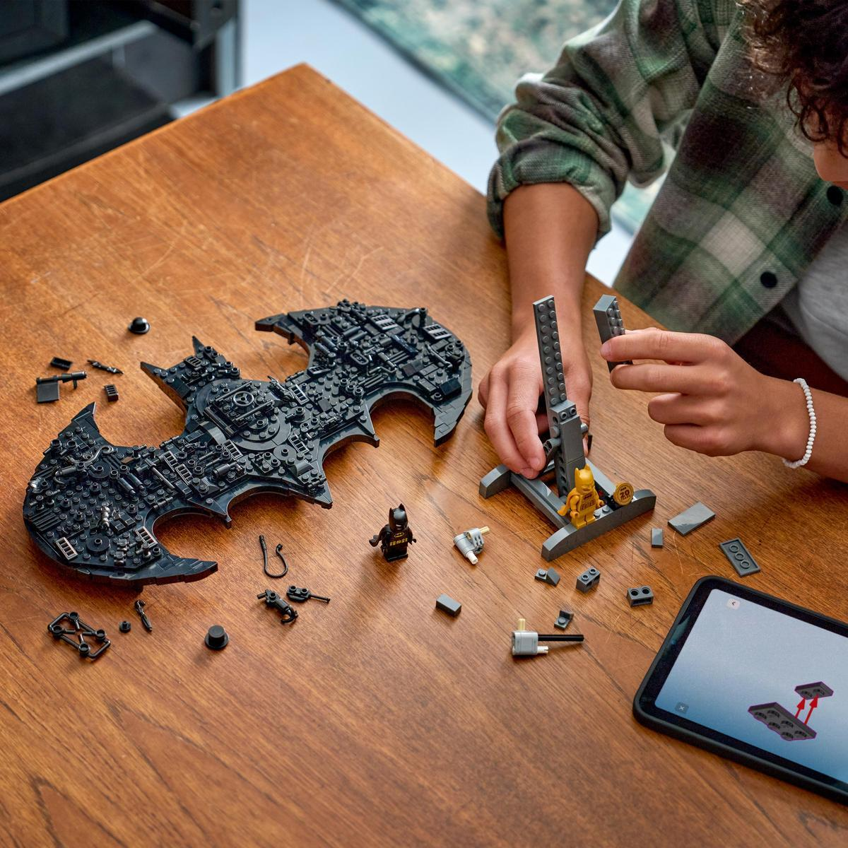 Bir kişi siyah Lego yarasa sembolü, figürinler ve tabletli bir stand inşa ediyor.