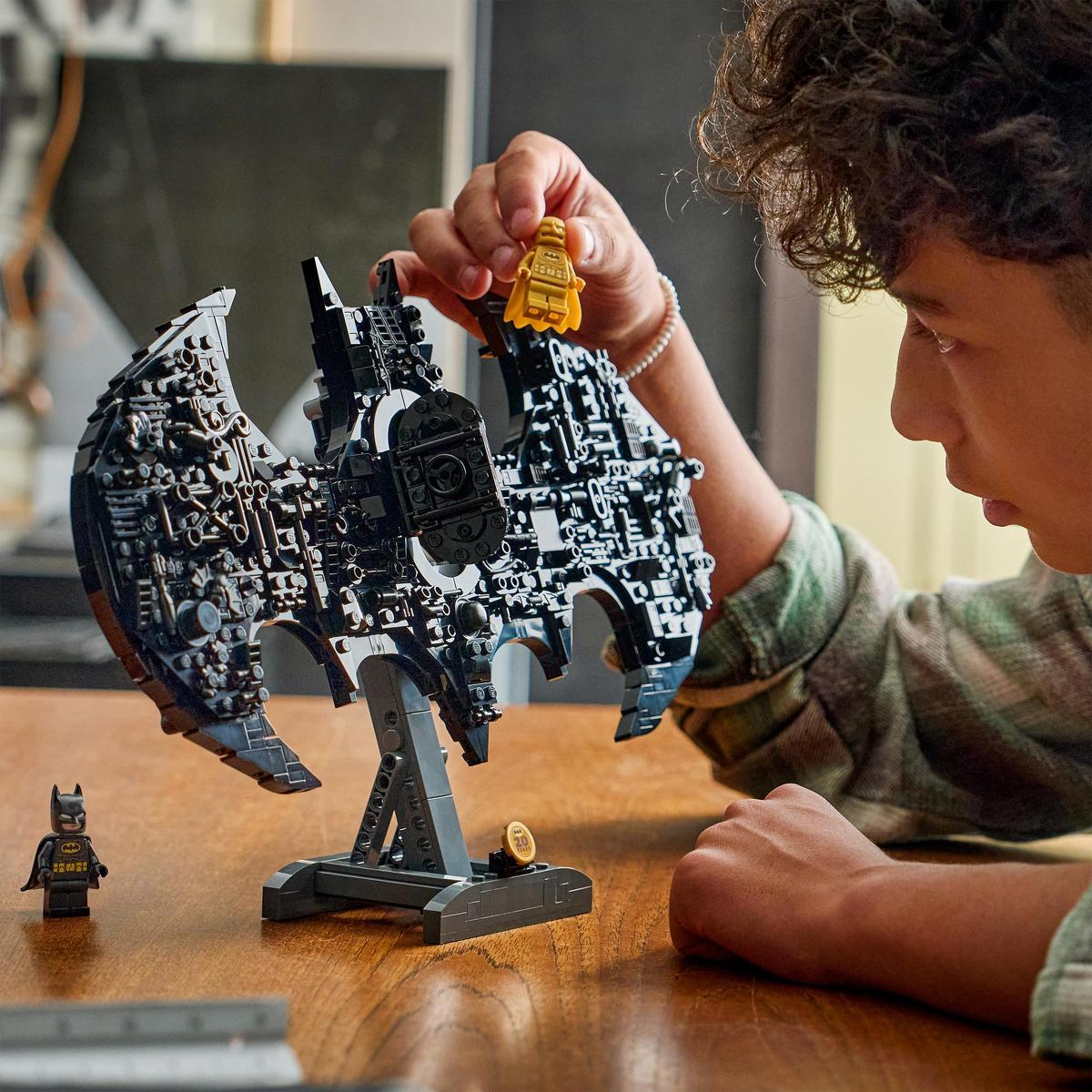 Bir kişi, sarı bir minifigür tutarak, bir stant üzerinde siyah bir Lego yarasa sembolü monte ediyor.