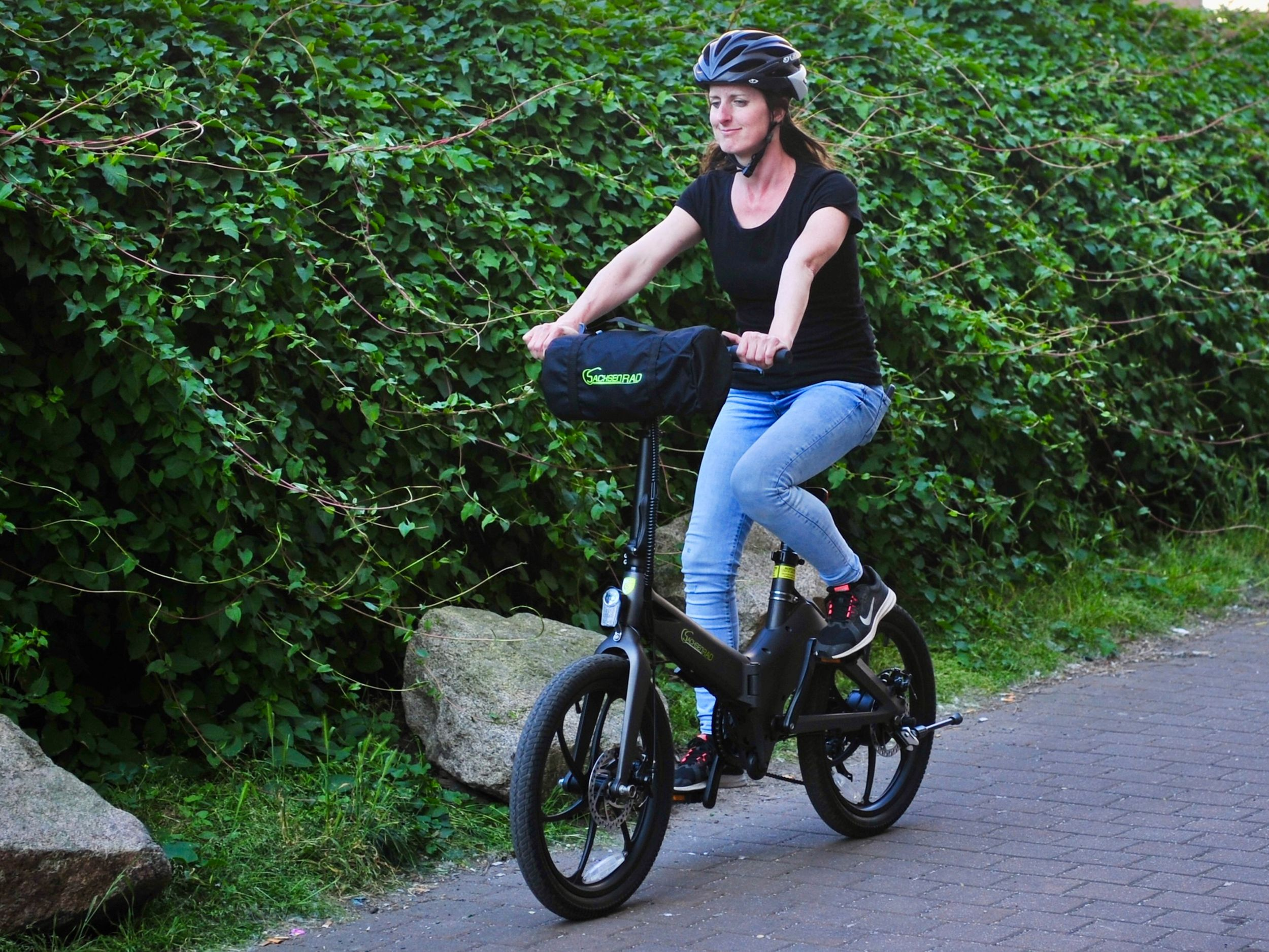Frau fährt E-Bike mit Helm, schwarzem Shirt und Jeans, vor grünem Blätterhintergrund.