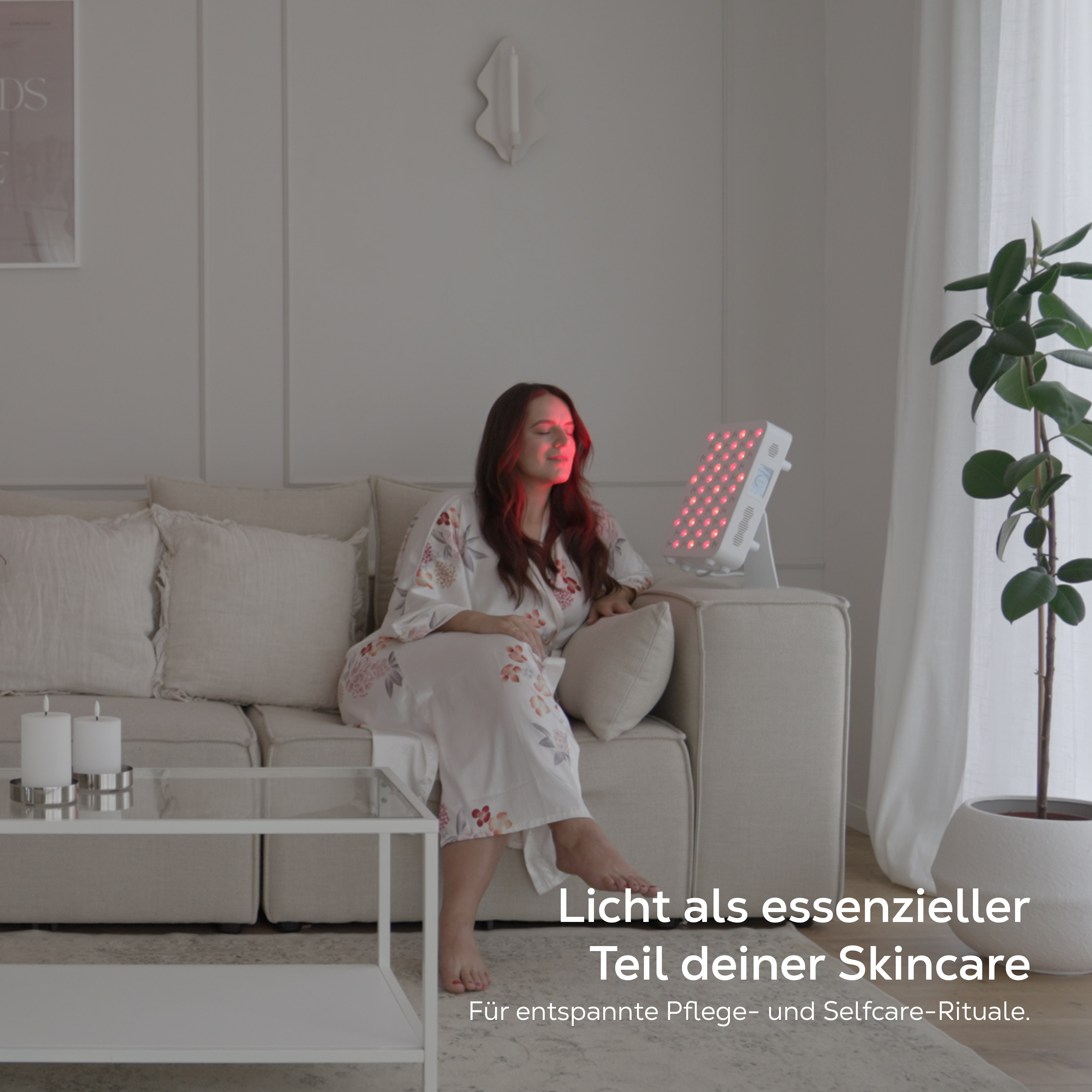 Frau sitzt auf Sofa, rotes Licht bestrahlt ihr Gesicht, Skincare-Gerät daneben.