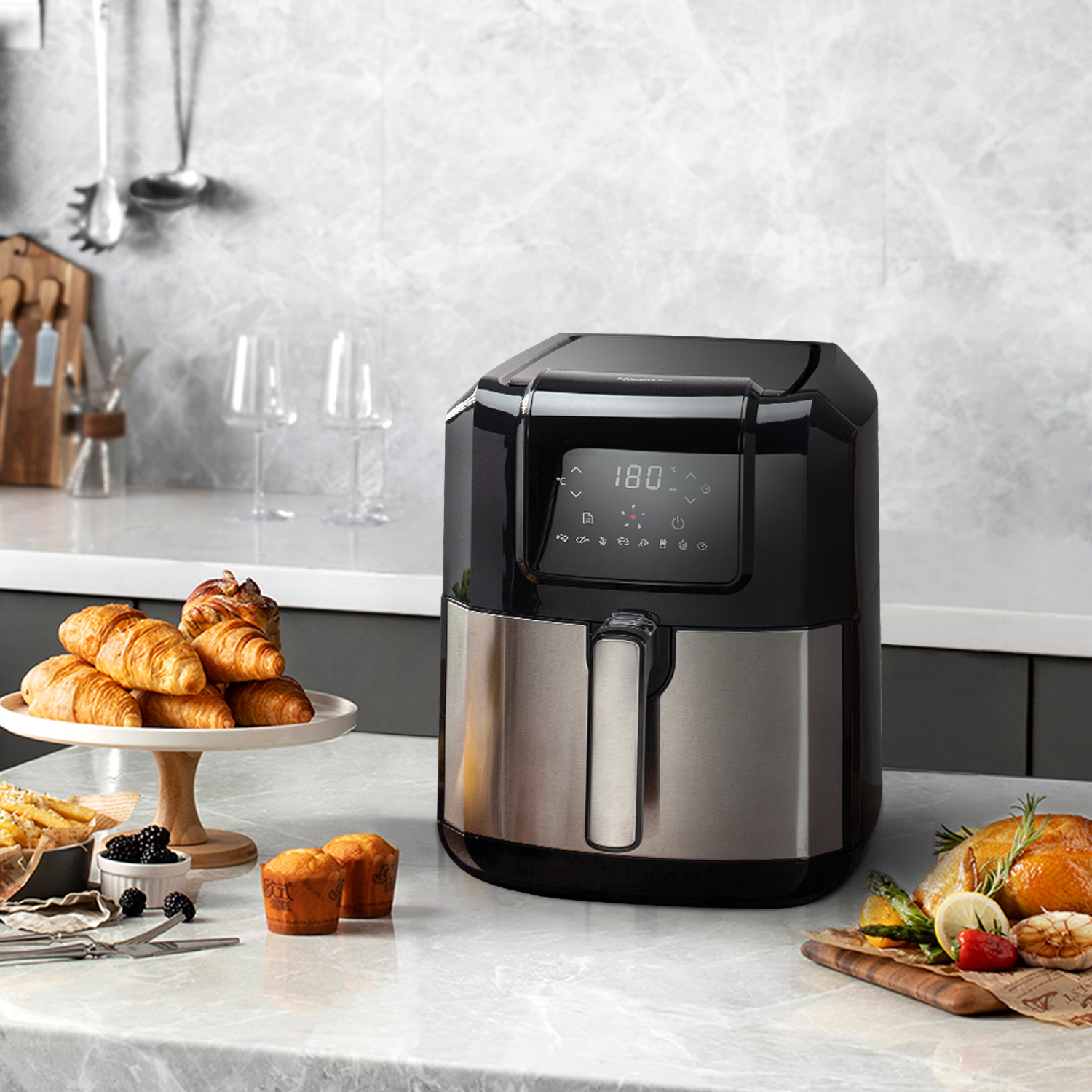 A black air fryer sits on a marble countertop with pastries and a roasted chicken.
