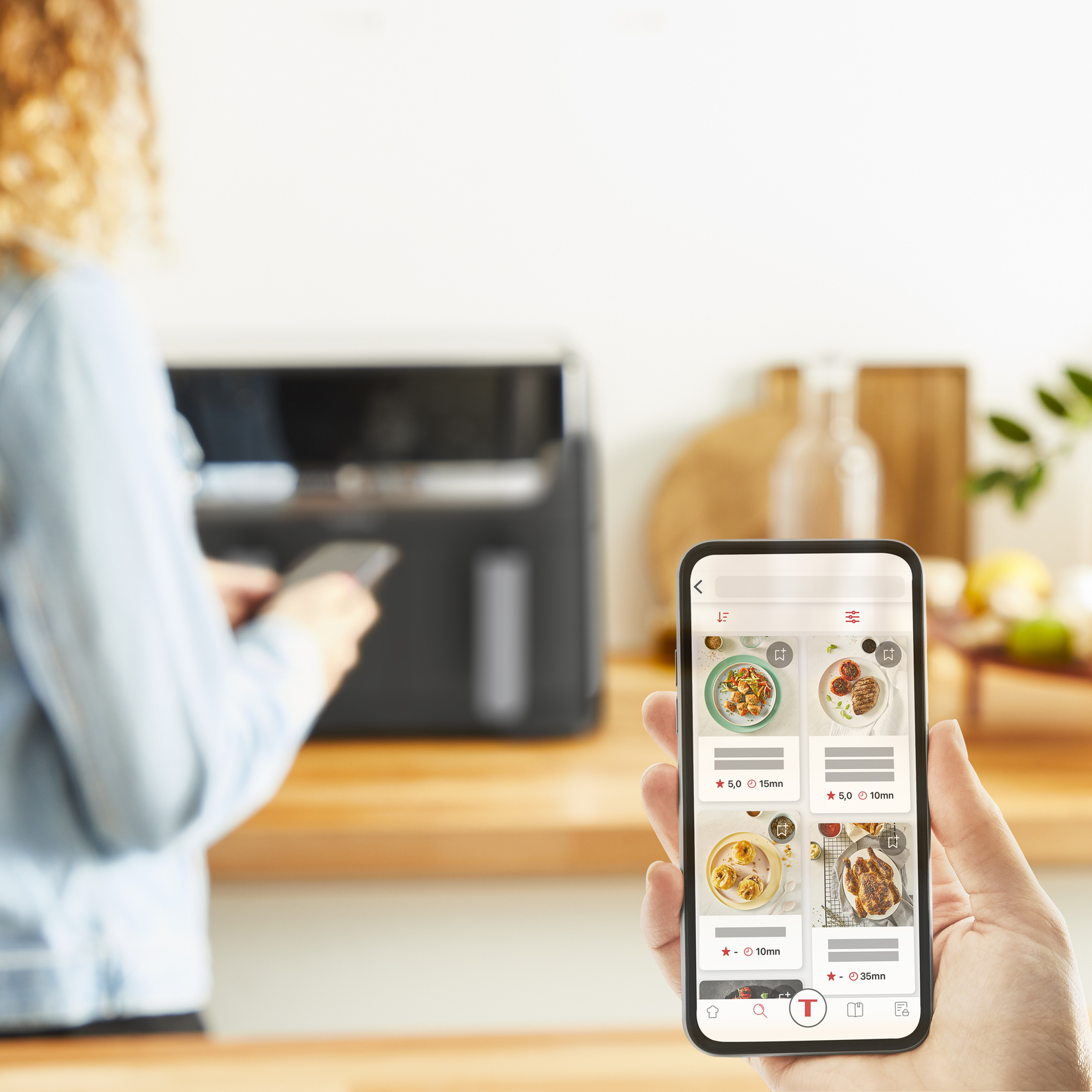 Osoba trzyma smartfon z aplikacją z przepisami, stojąc obok frytkownicy beztłuszczowej.