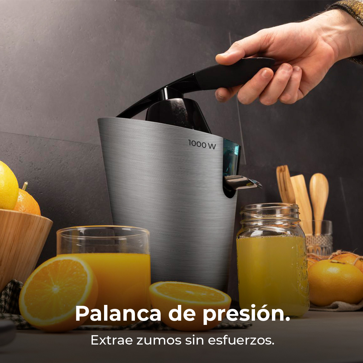 Una mano acciona un exprimidor plateado junto a naranjas y vasos de zumo.