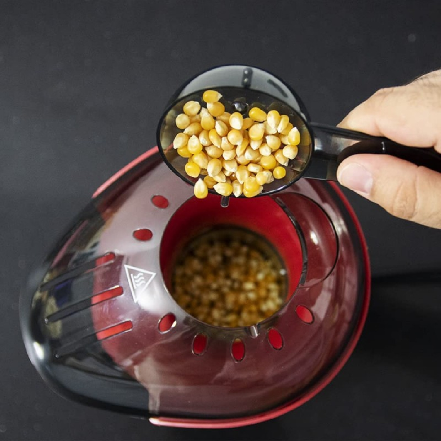 Una mano versa chicchi di mais per popcorn da un cucchiaio in una macchina per popcorn rossa e trasparente.