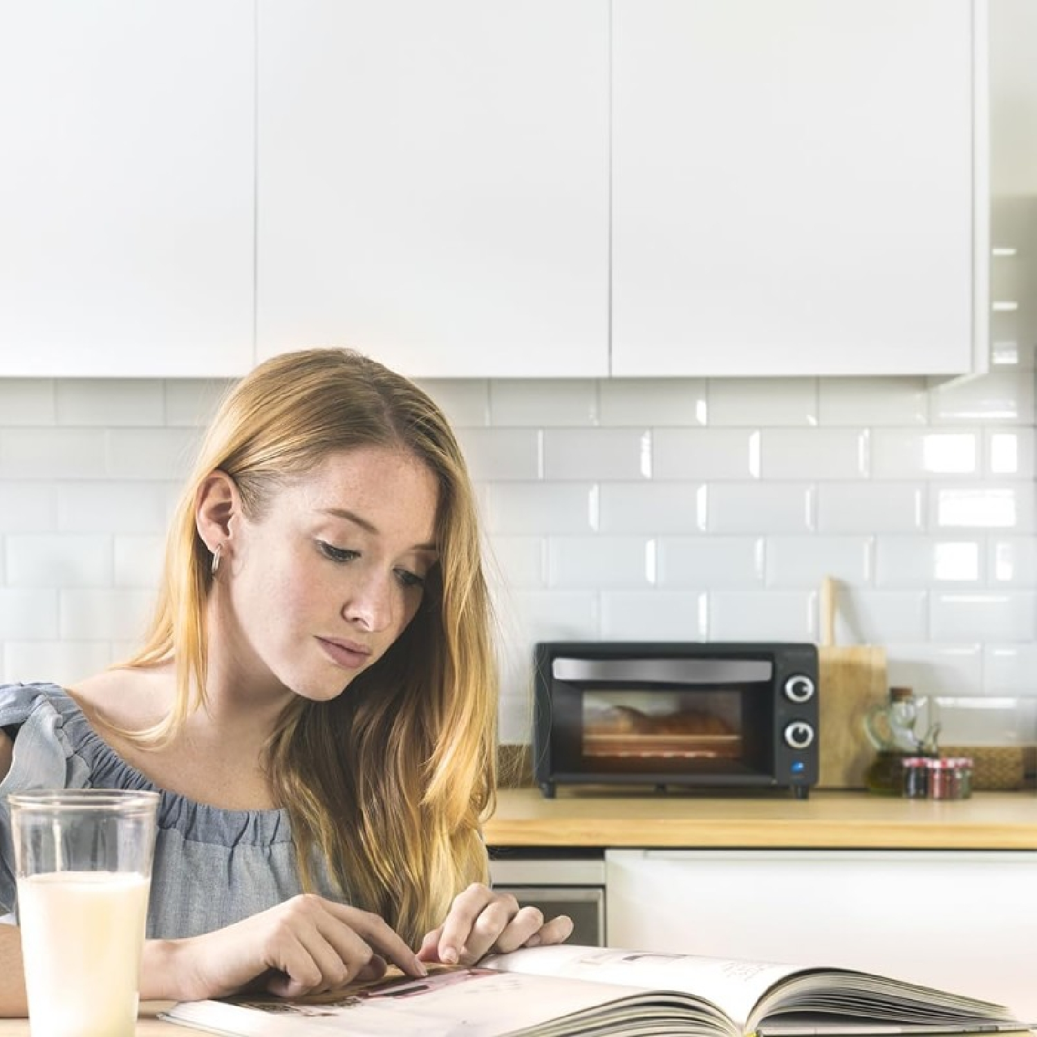 Una joven pelirroja lee un libro en una cocina. A su lado hay un vaso de leche.