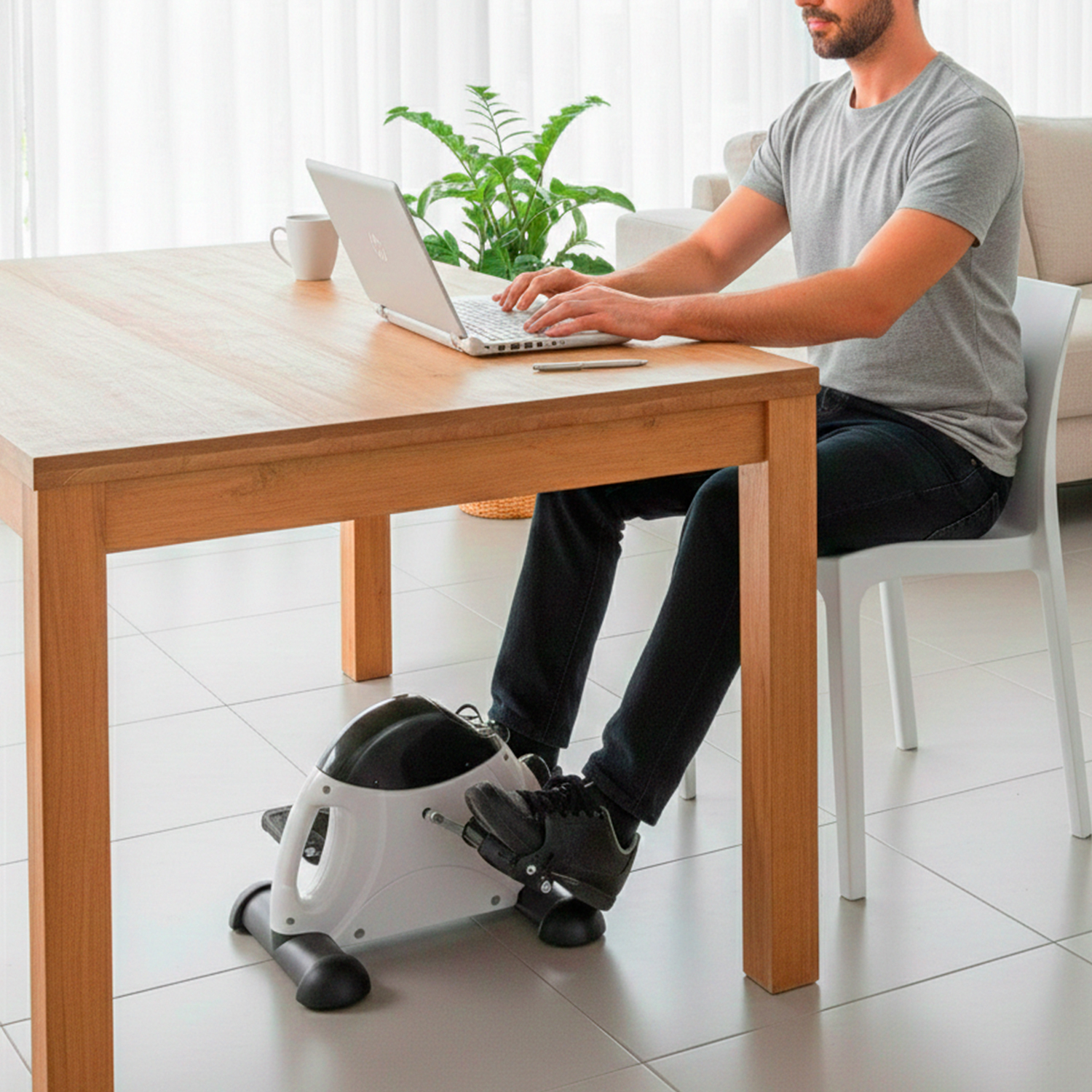 Hombre pedalea mini bicicleta estática mientras trabaja en laptop.