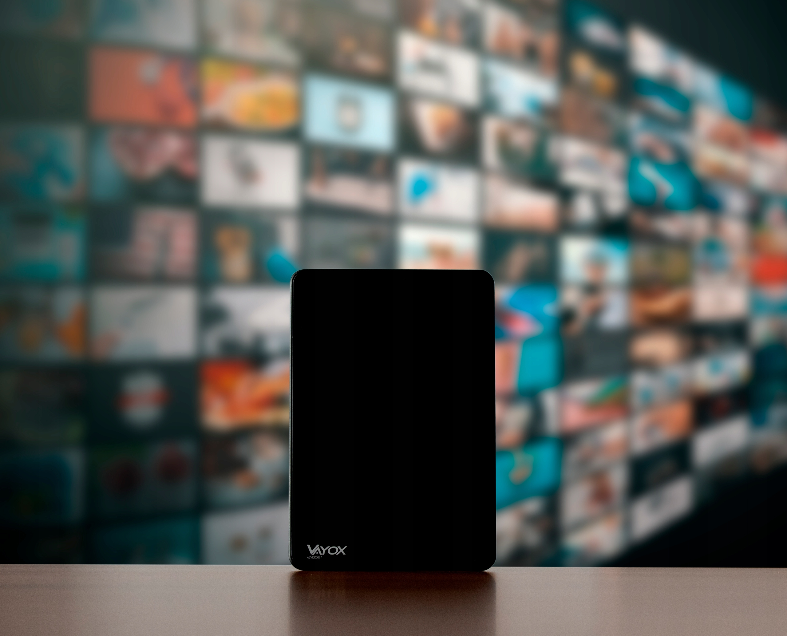 Czarne urządzenie z logo VAYOX stoi na drewnianym stole. W tle wiele nieostrych ekranów.