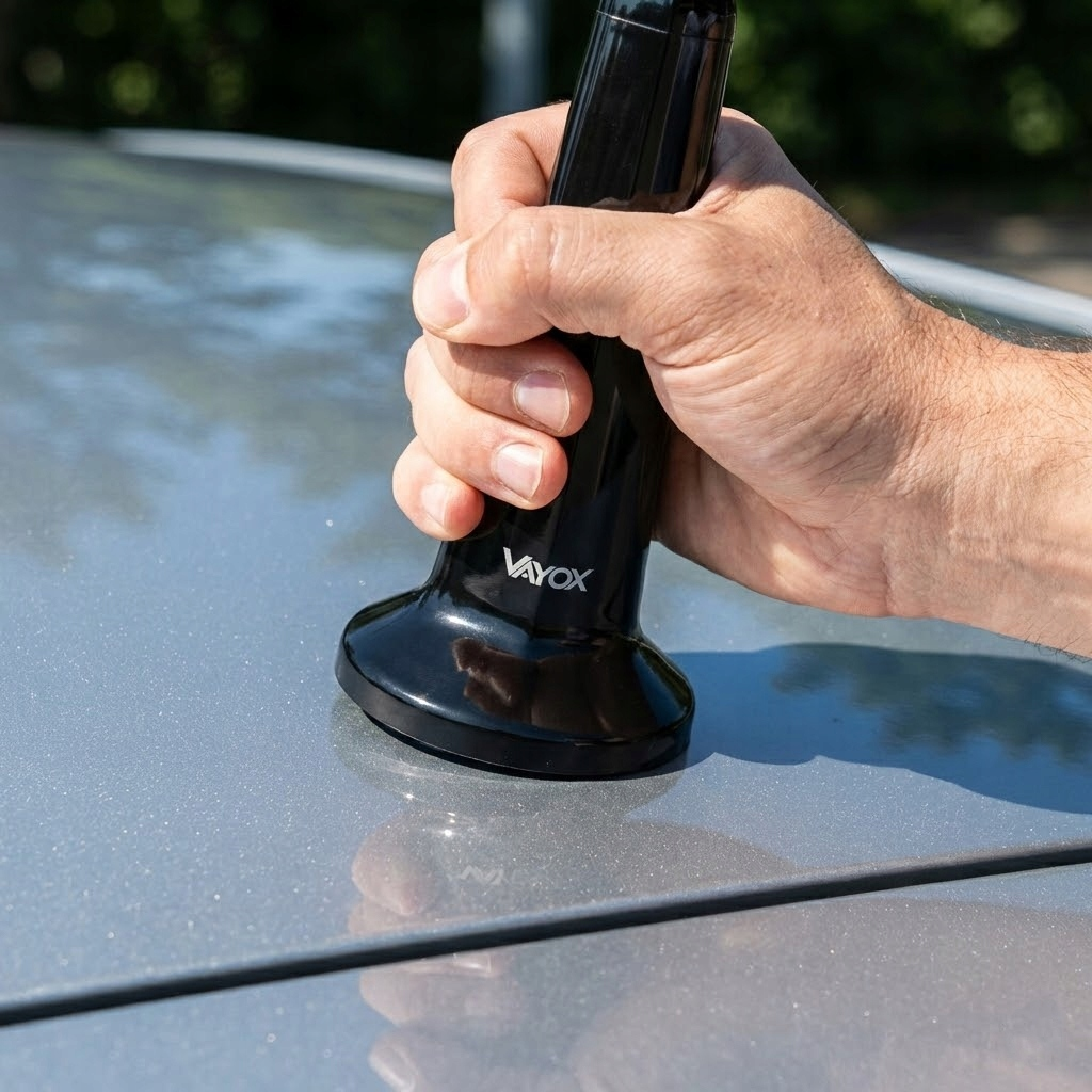 Ręka trzyma czarne urządzenie Vayox na szarym samochodzie.