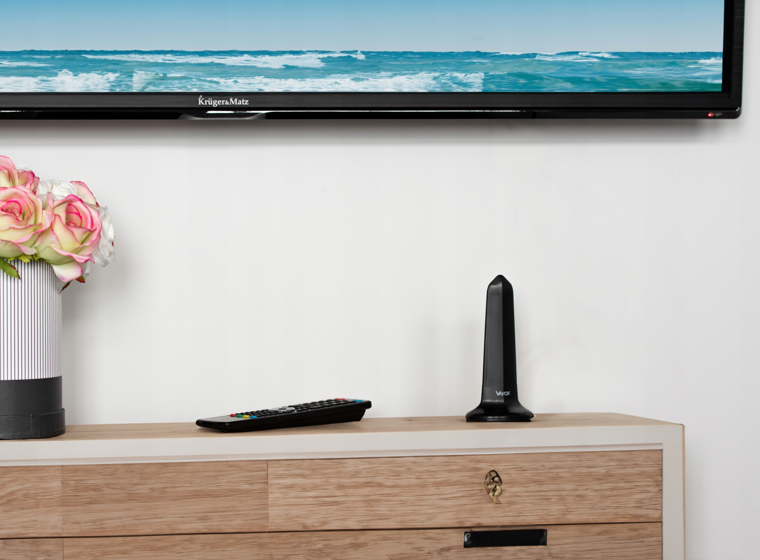 Czarny odbiornik pilota stoi na drewnianej szafce obok pilota i kwiatów.