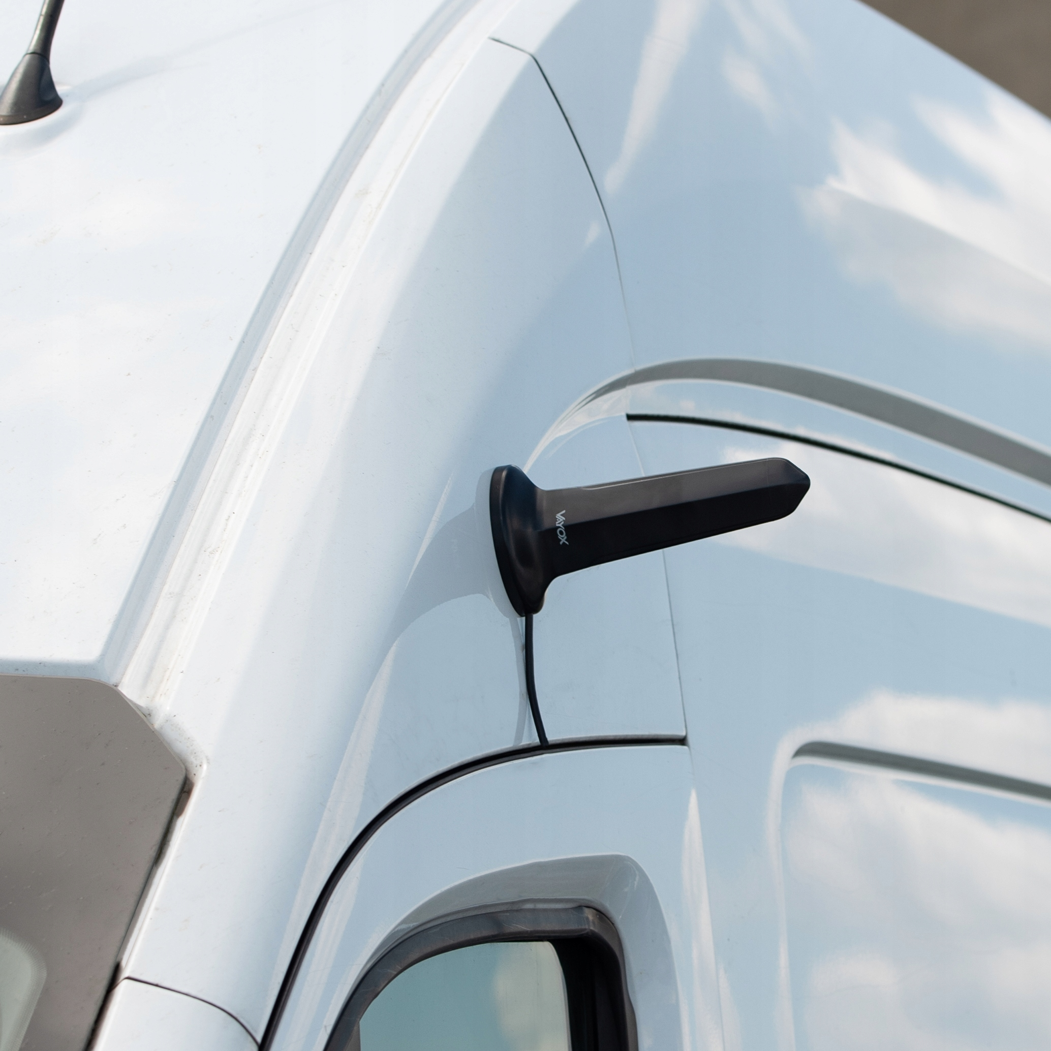 Czarna podstawa anteny VAUX na białym dachu pojazdu z odbiciem nieba.