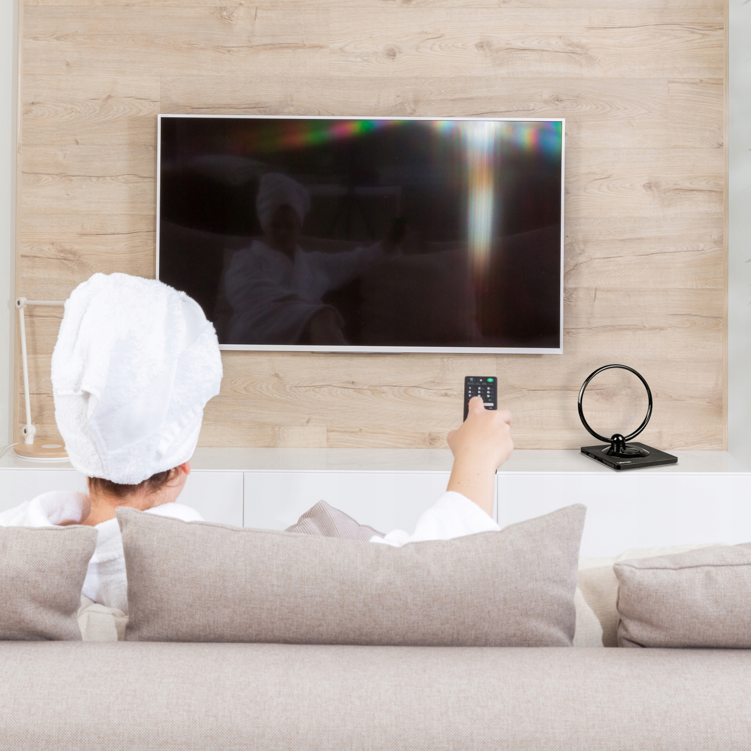 Kobieta w szlafroku i ręczniku ogląda TV, trzymając pilota.