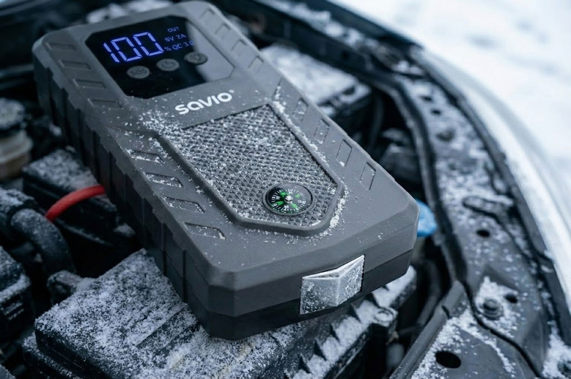 Szary power bank z wyświetlaczem cyfrowym na zaśnieżonym silniku auta.