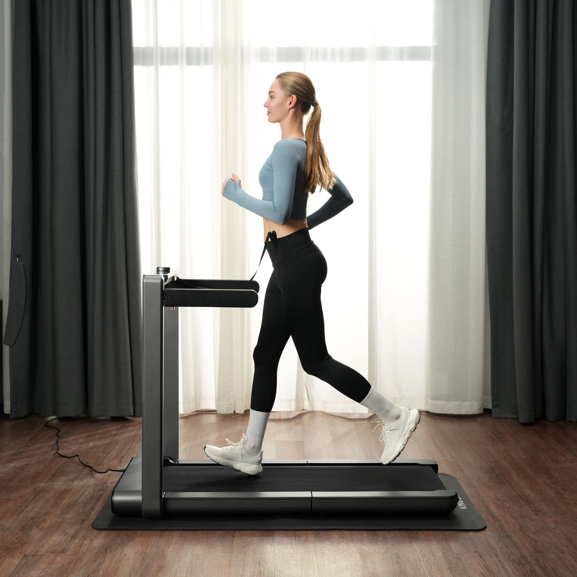 Una mujer corre en una cinta de correr en una habitación con cortinas y ventana.