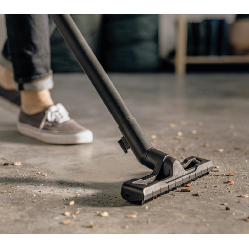 Głowica odkurzacza czyści brudną podłogę betonową, osoba w tle nosi trampki.