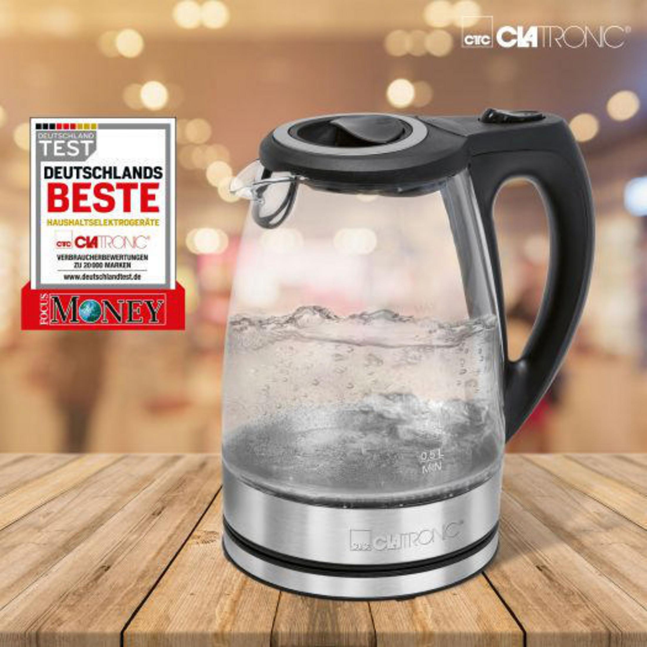 Szklany czajnik elektryczny z srebrną podstawą i czarnym uchwytem stoi na drewnianym stole.