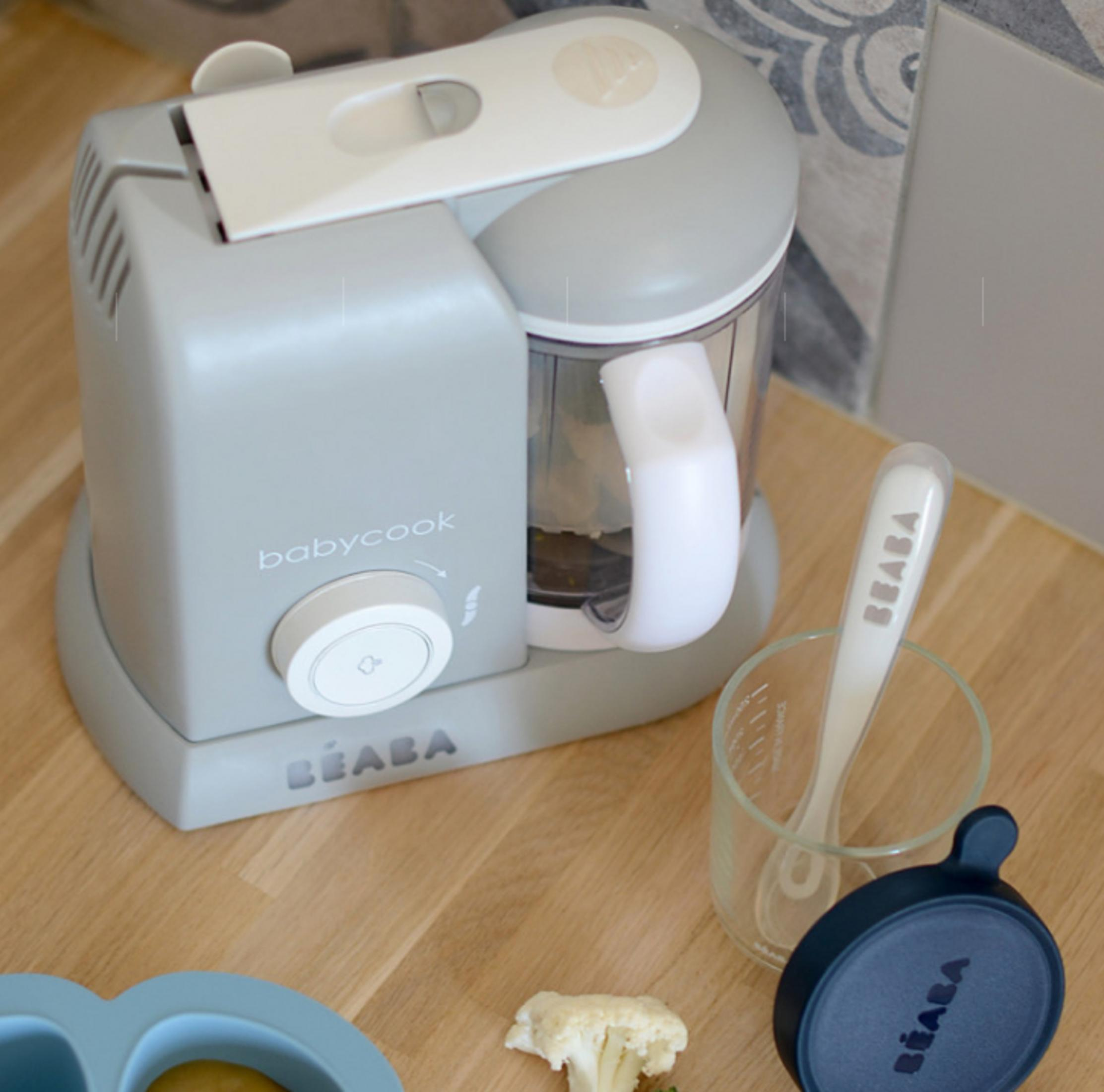 Grauer Babycook-Mixer mit Löffel, Glas und Deckel auf Holztisch.