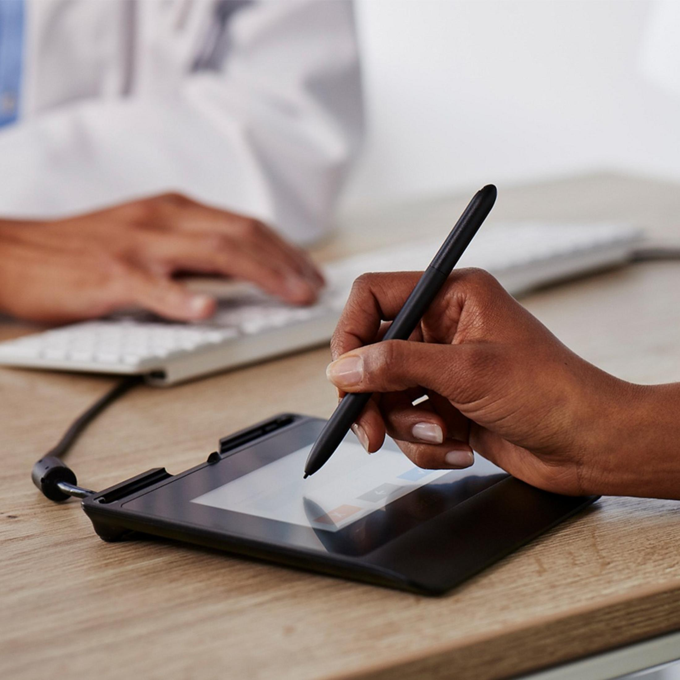 Mano con penna firma su tablet, uomo digita tastiera sullo sfondo.