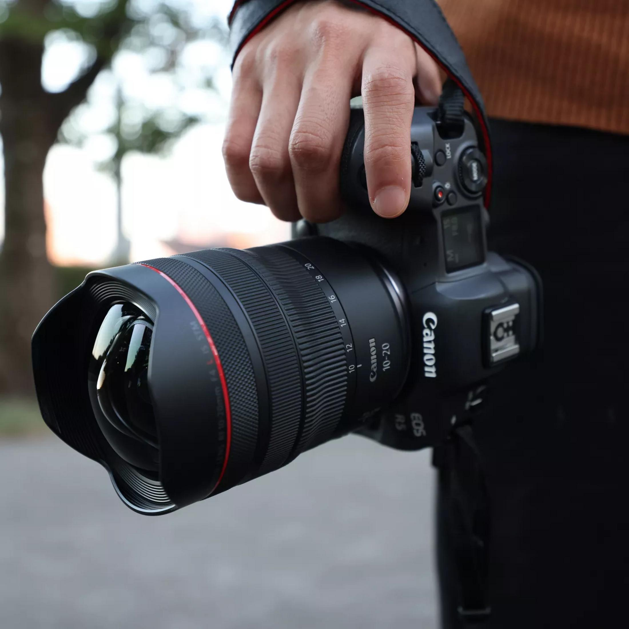 Een hand houdt een zwarte Canon EOS R5 camera met een grote lens vast.