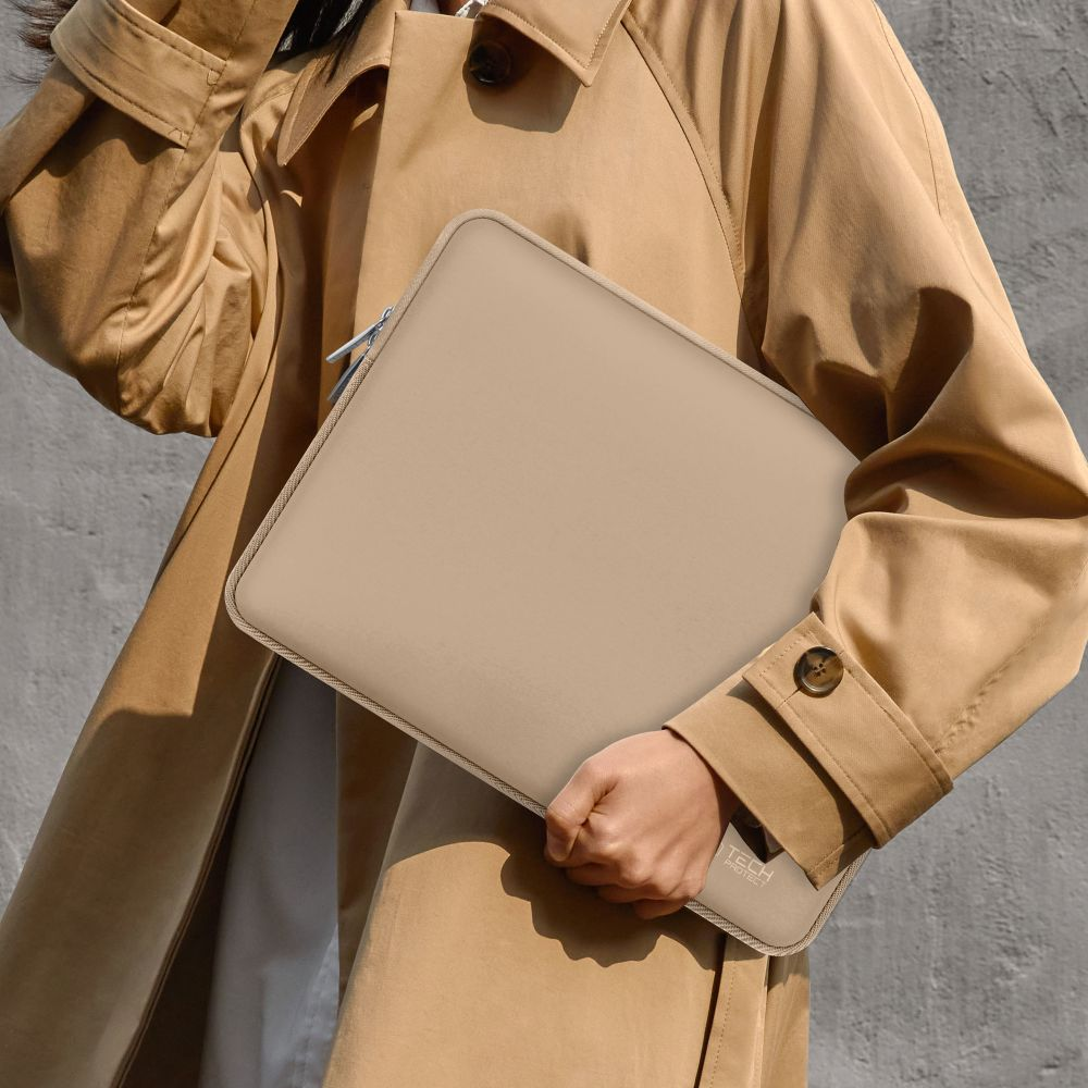 Eine Person in einem beigen Mantel hält eine beige Laptoptasche. Grauer Hintergrund.