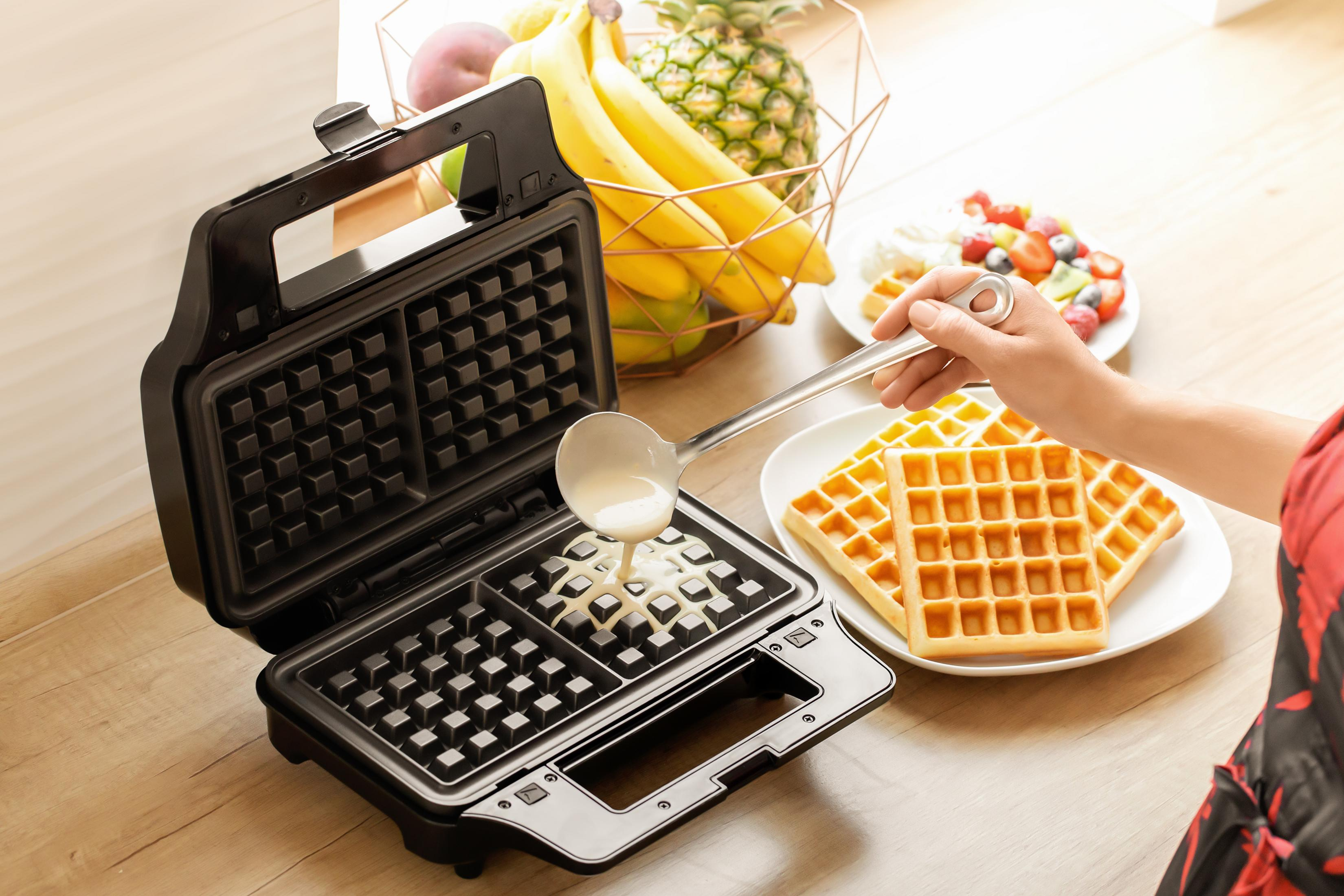 Flüssiger Teig wird in eine offene Waffelform gegossen. Daneben liegen fertige Waffeln.