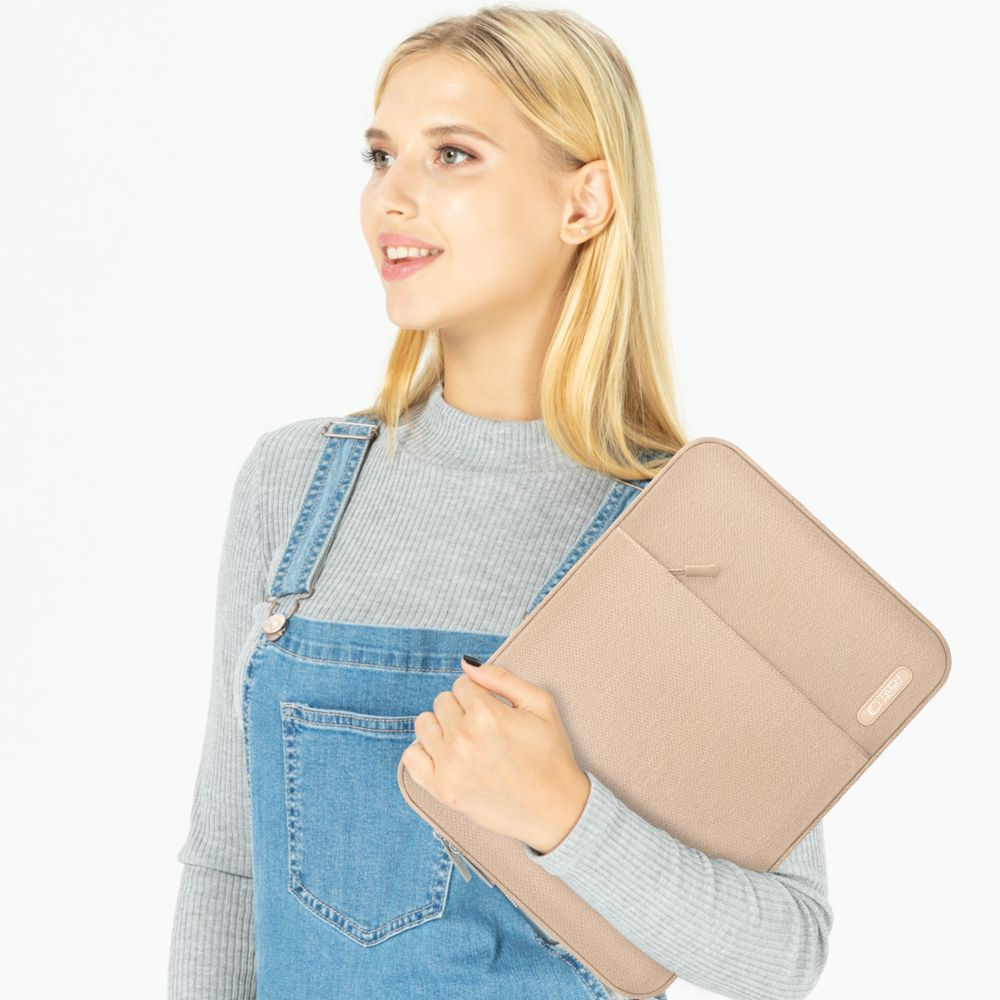 Eine blonde Frau in Latzhose und grauem Pullover hält eine hellbraune Laptoptasche.