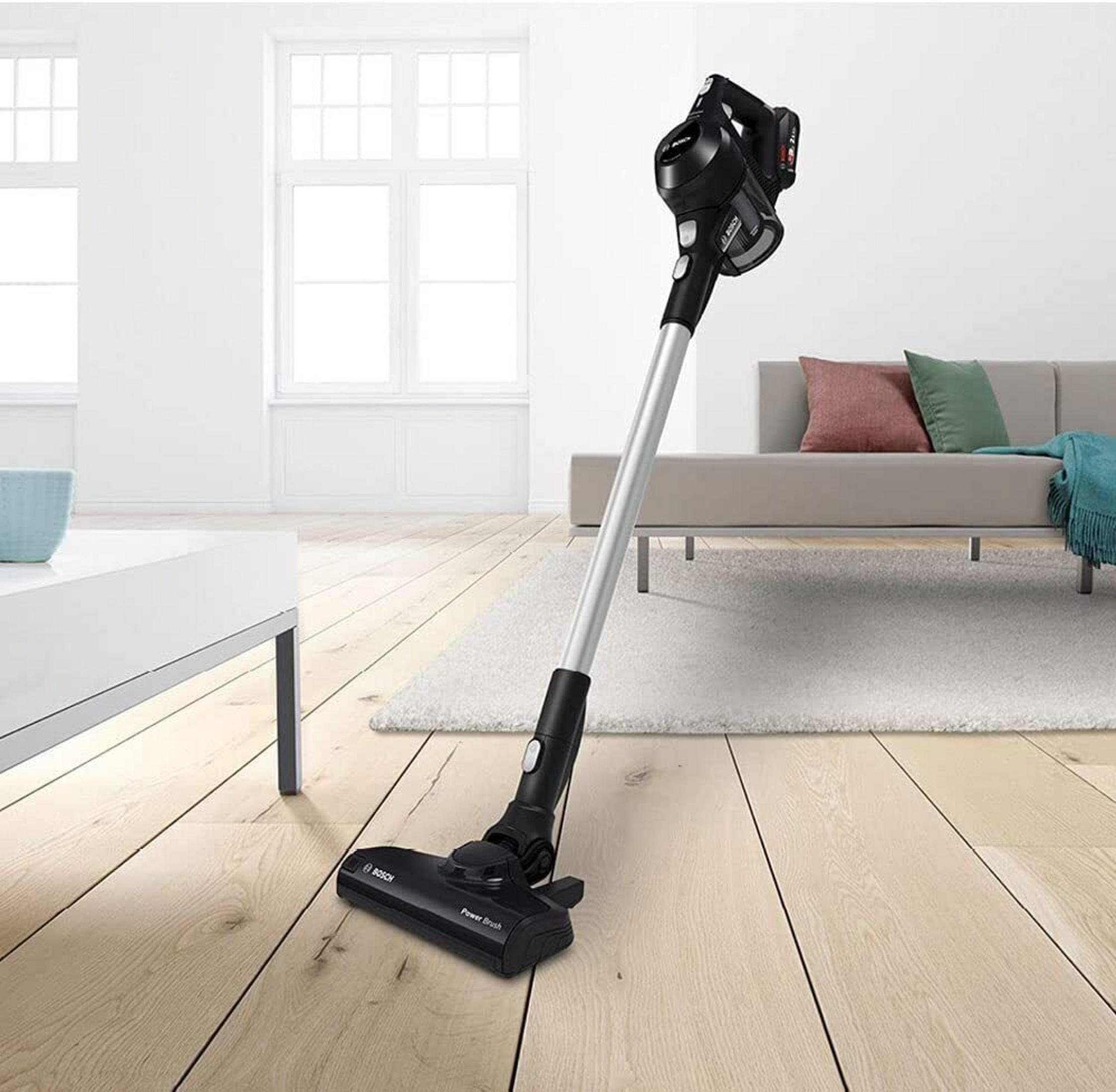 Aspiradora inalámbrica Bosch negra sobre suelo de madera clara, sofá al fondo.