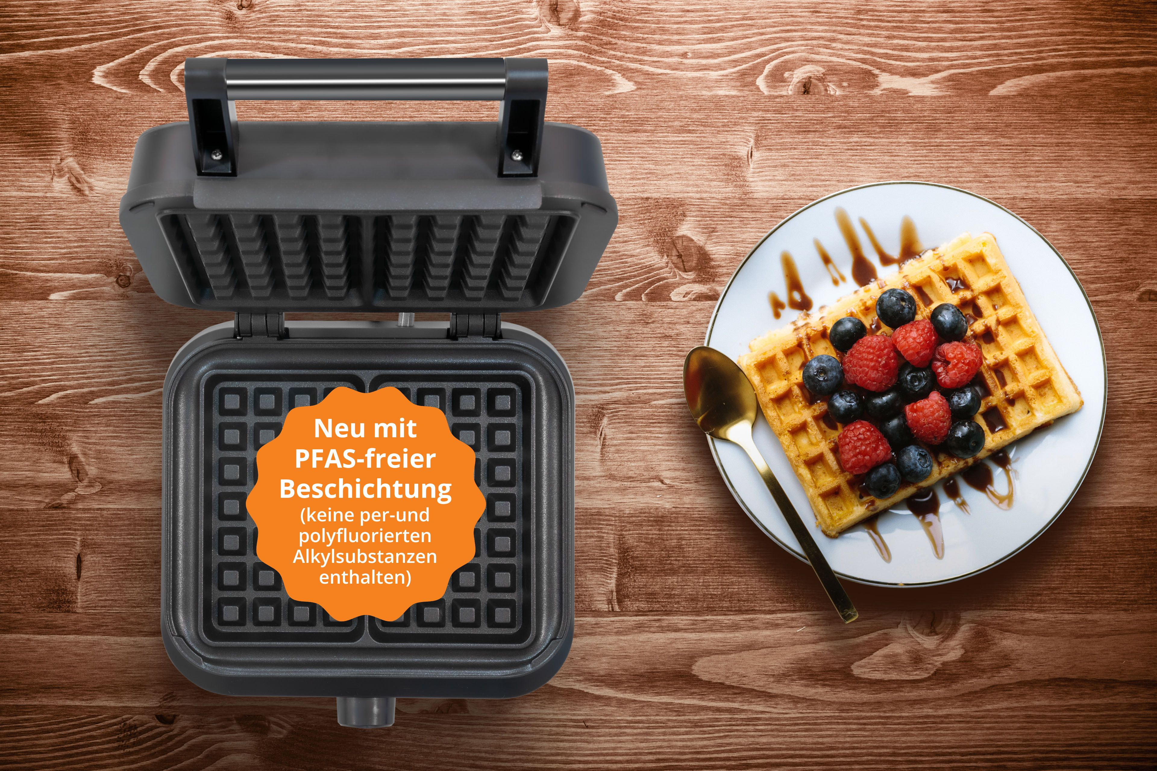 Ein geöffneter Waffelautomat neben einem Teller mit Waffel, Beeren und Sirup.