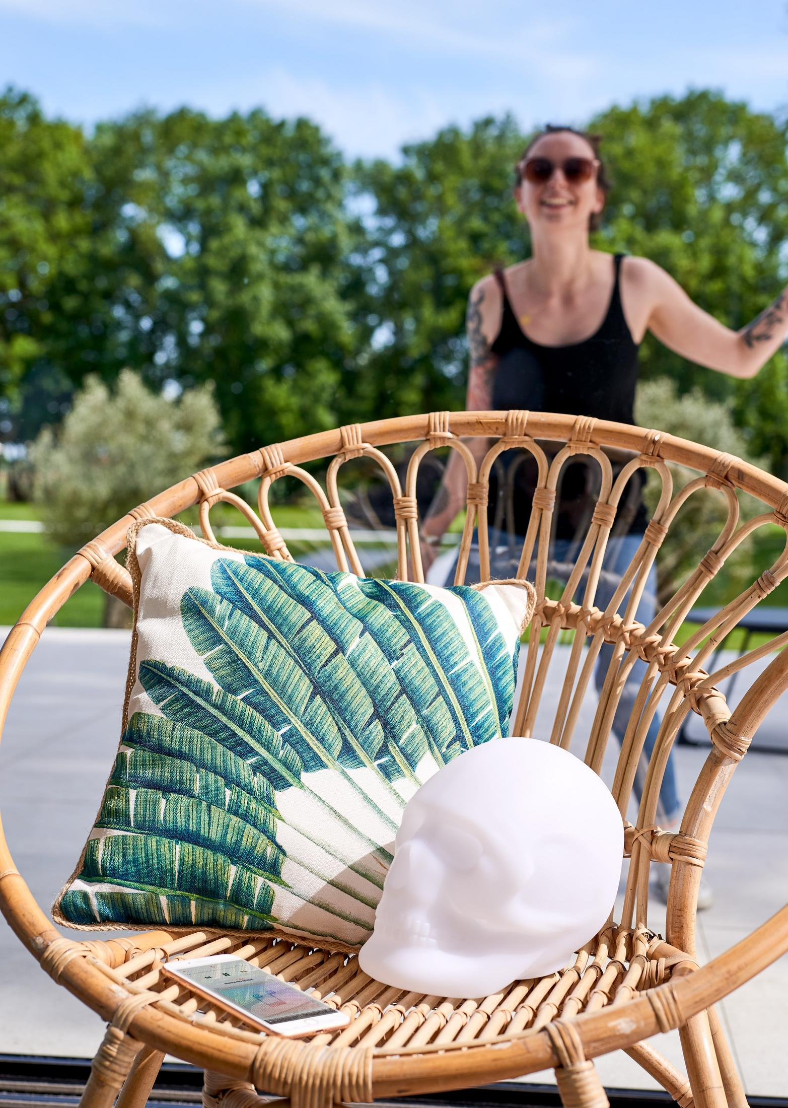 Ein Rattanstuhl mit Kissen, Handy und Totenkopf-Lampe. Eine Frau ist unscharf im Hintergrund.