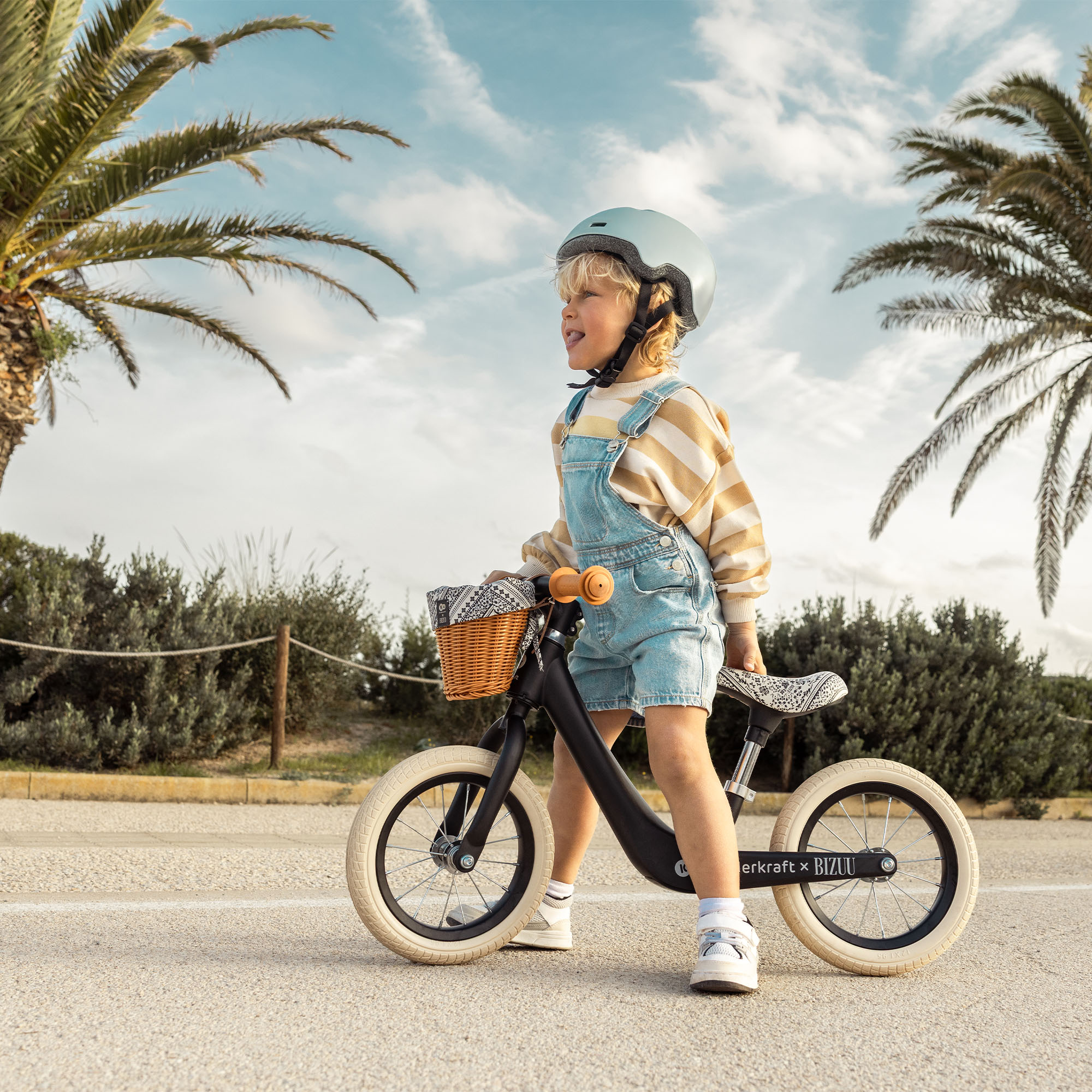 Ein Junge mit Helm und Latzhose auf einem Laufrad vor Palmen und blauem Himmel.