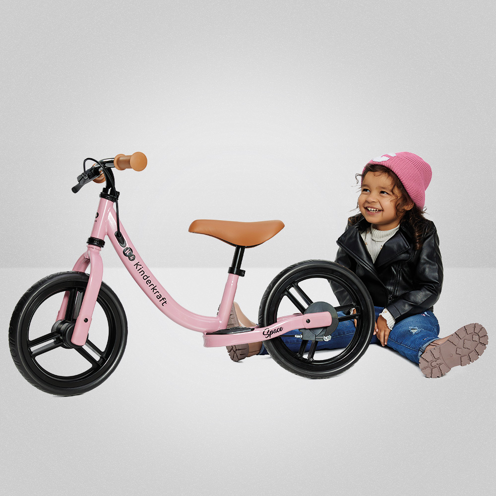 Ein rosa Laufrad steht neben einem lächelnden Mädchen in Lederjacke und Mütze.