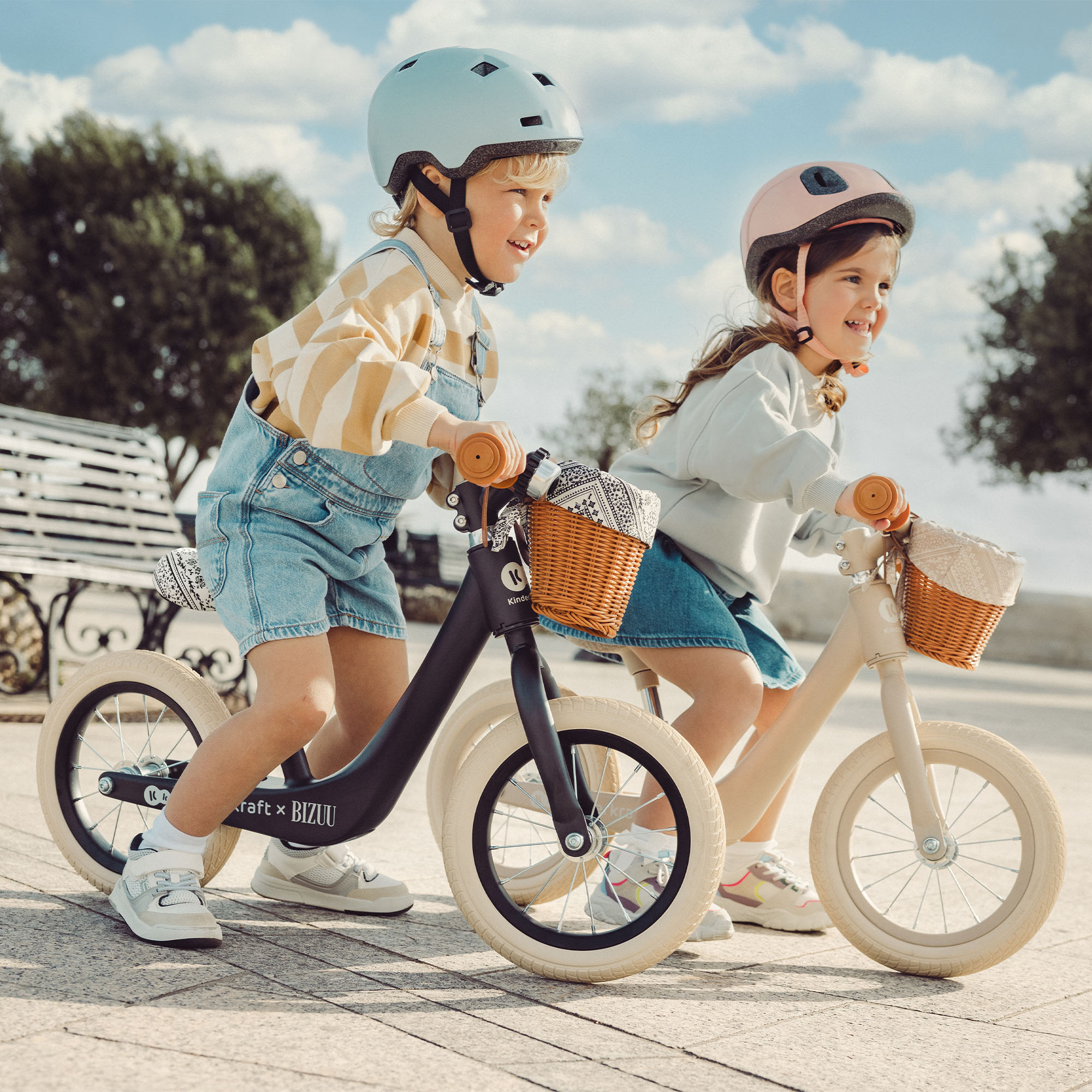 Zwei Kinder fahren auf Laufrädern mit Körben vorne, im Freien.
