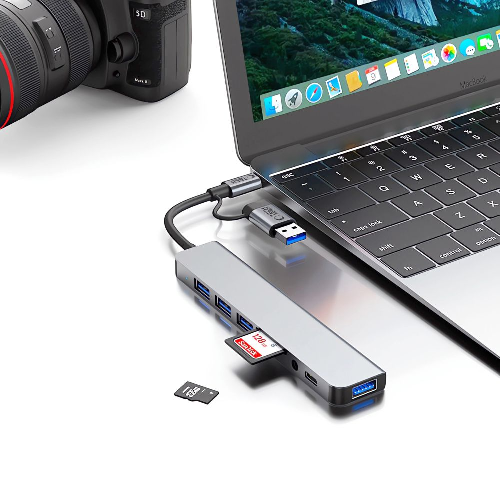 Un hub USB-C con scheda SD è collegato a un laptop. Una fotocamera è sullo sfondo.