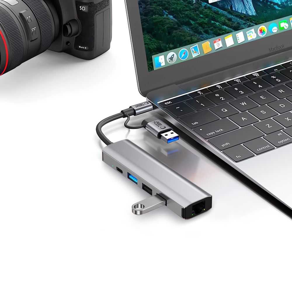 Un hub USB argentato si collega a un MacBook, con una fotocamera sullo sfondo.