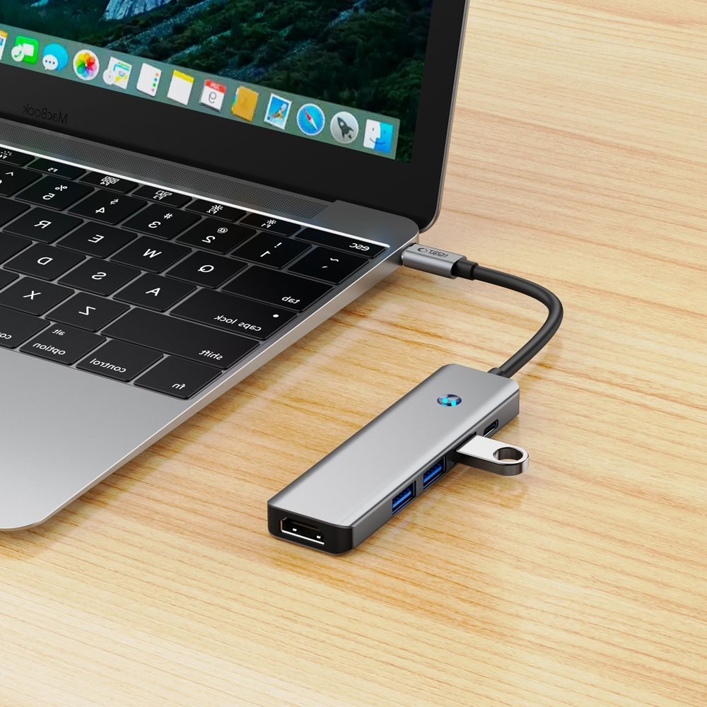 Un hub USB-C grigio con porte HDMI e USB, collegato a un MacBook su una scrivania di legno.