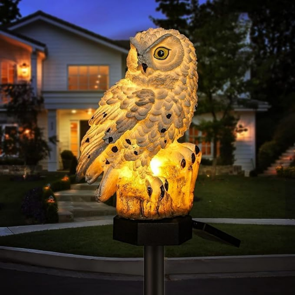 Beleuchtete Eulenfigur auf einem Baumstumpf im Vorgarten vor einem Haus bei Nacht.