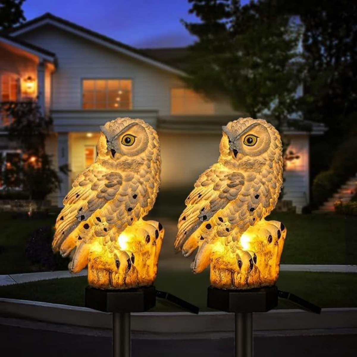 Zwei leuchtende Eulen-Gartenleuchten stehen vor einem Haus im Abendlicht.