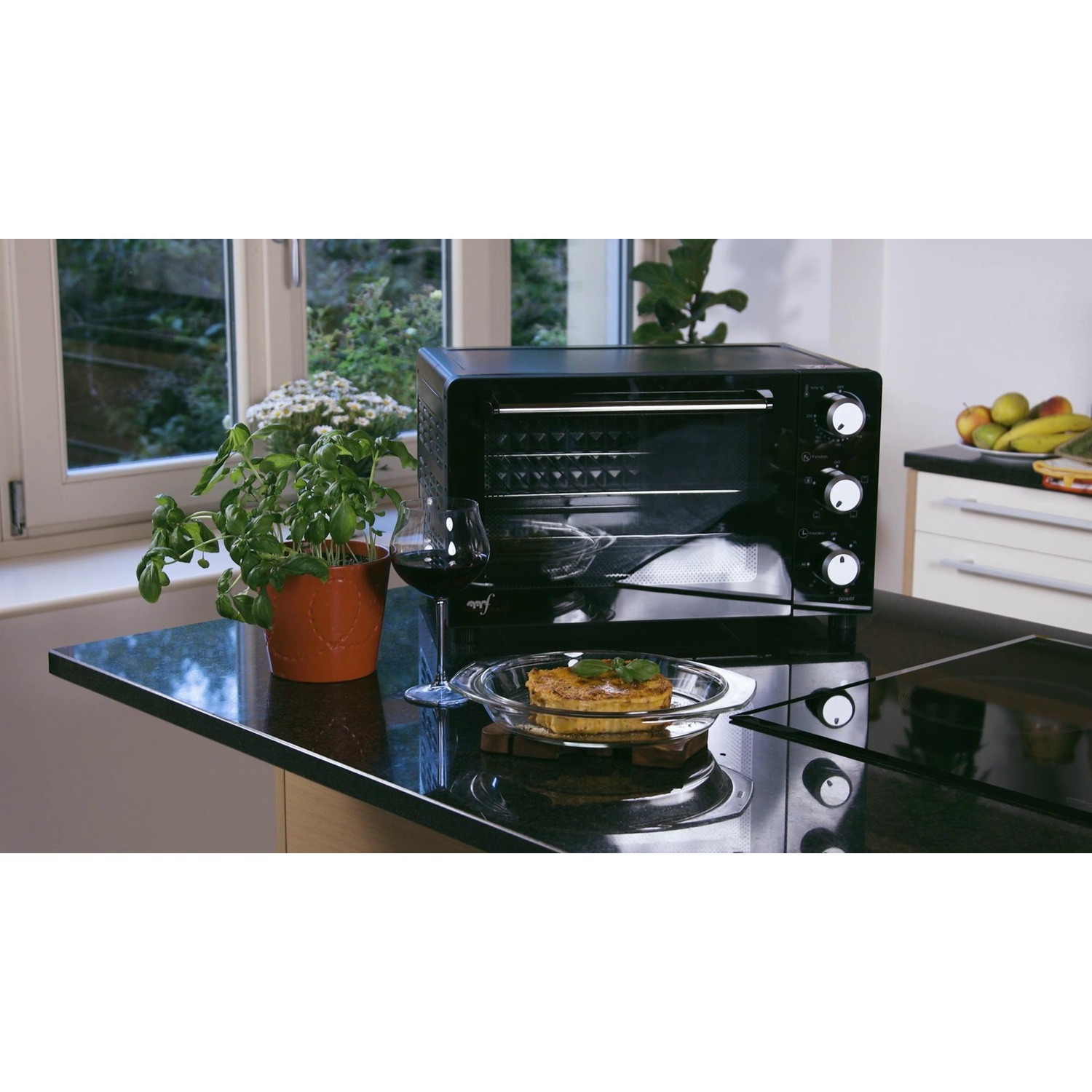 Czarny mini piekarnik na blacie kuchennym obok kieliszka czerwonego wina i potrawy.